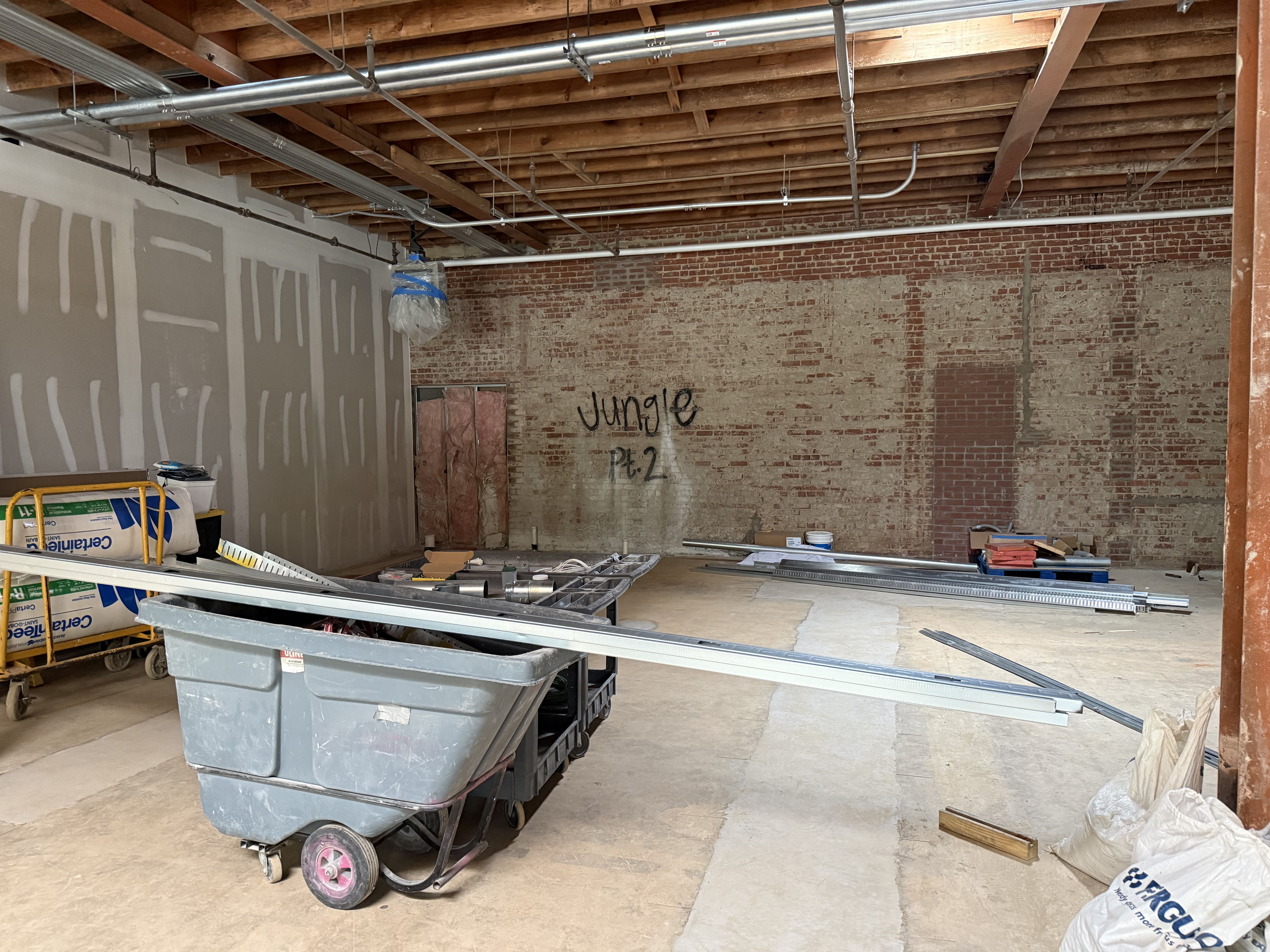 Unfinished industrial interior with exposed wooden beams and overhead metal pipes. Brick back wall features graffiti reading "Jungle". A large gray dumpster sits amid scattered construction debris and drywall sheets.