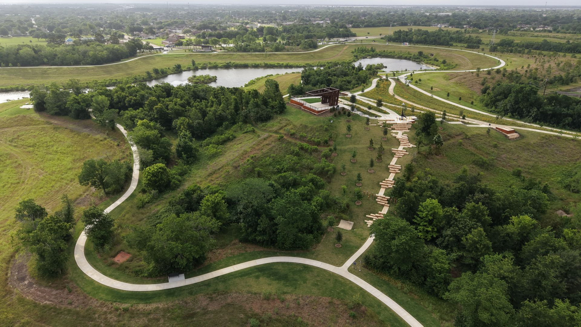 Aerial view of a park with winding white paths, a central pond, dense trees, a wooden pavilion on a hill, and stepped terraces leading to green fields.