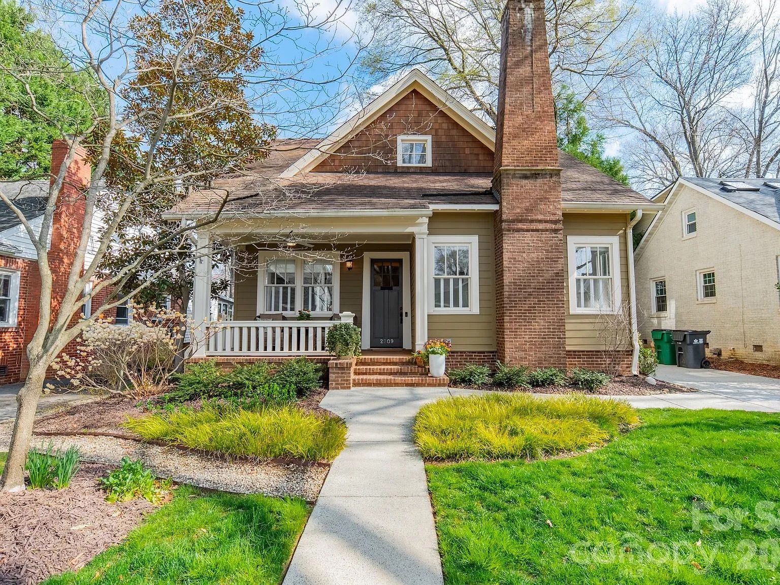 Front view of a cozy olive-siding house with a tall brick chimney, white trim, and a small porch. Green lawn, ornamental grasses, and a leafless tree frame the scene under a blue sky.