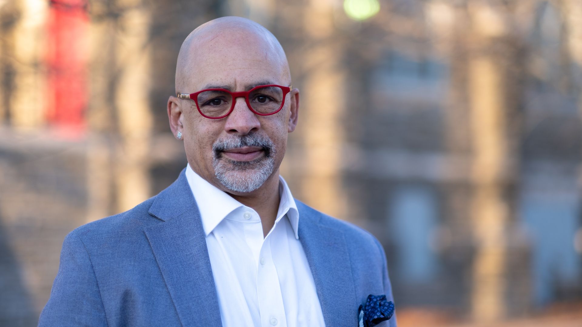 A bald man with a gray goatee wearing red eyeglasses, a light blue blazer, and a white shirt, standing outdoors with a softly blurred background.