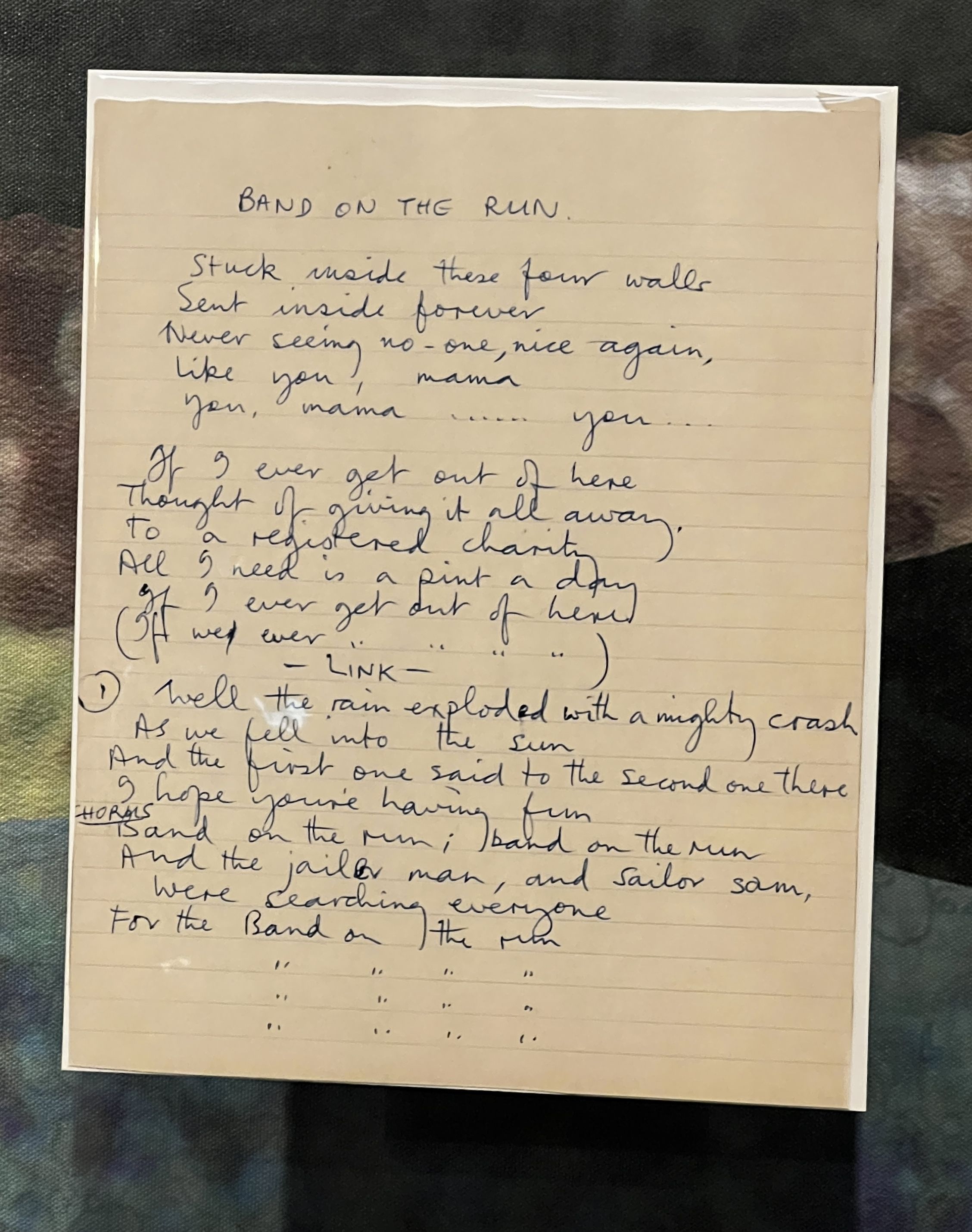 Handwritten lyric sheet titled "Band on the Run" in blue ink on lined beige paper, inside a clear plastic sleeve, resting on a dark, patterned fabric background.