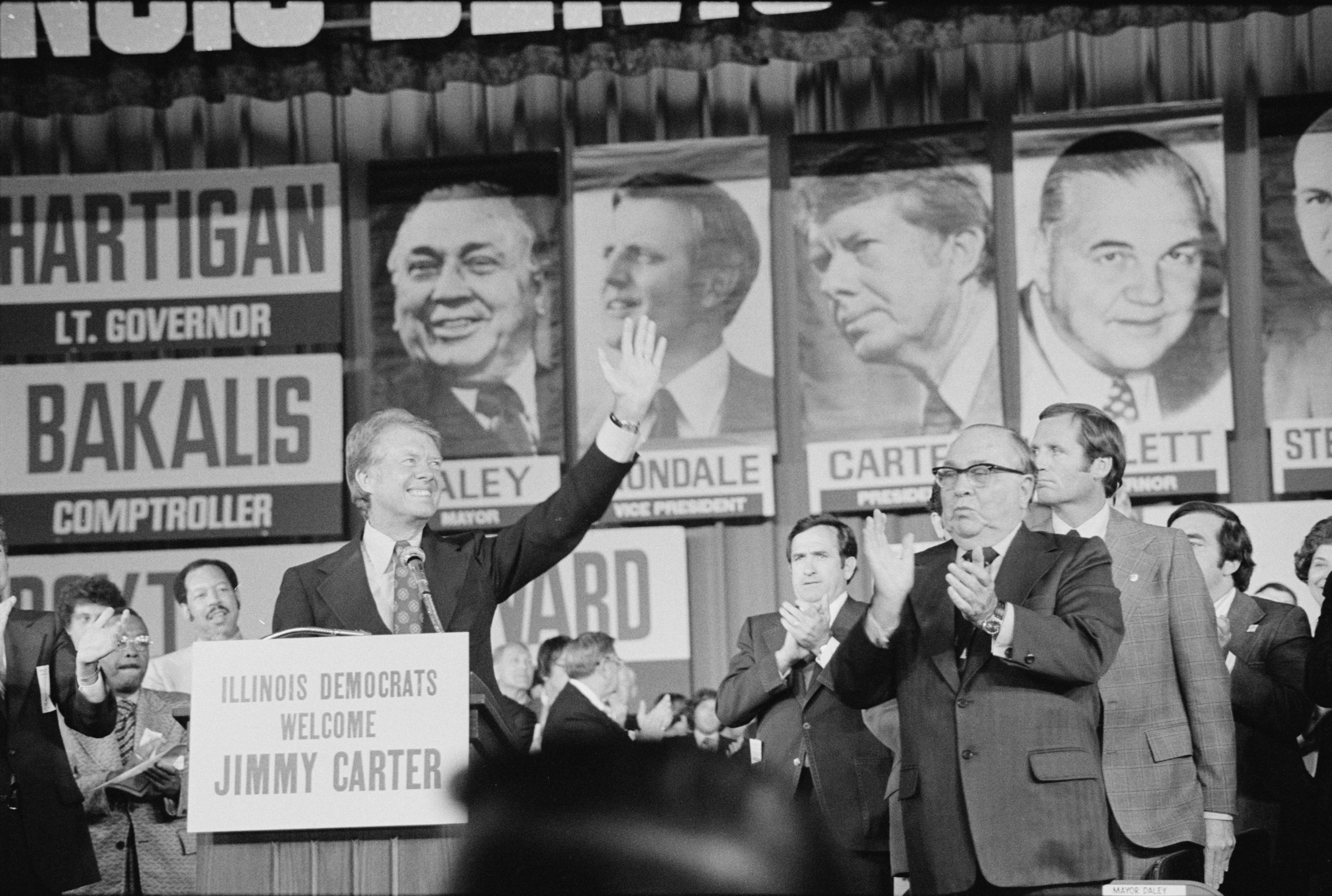black and white photo of Jimmy Carter standing on a stage and waving next to Chicago mayor Richard Daley in 1976