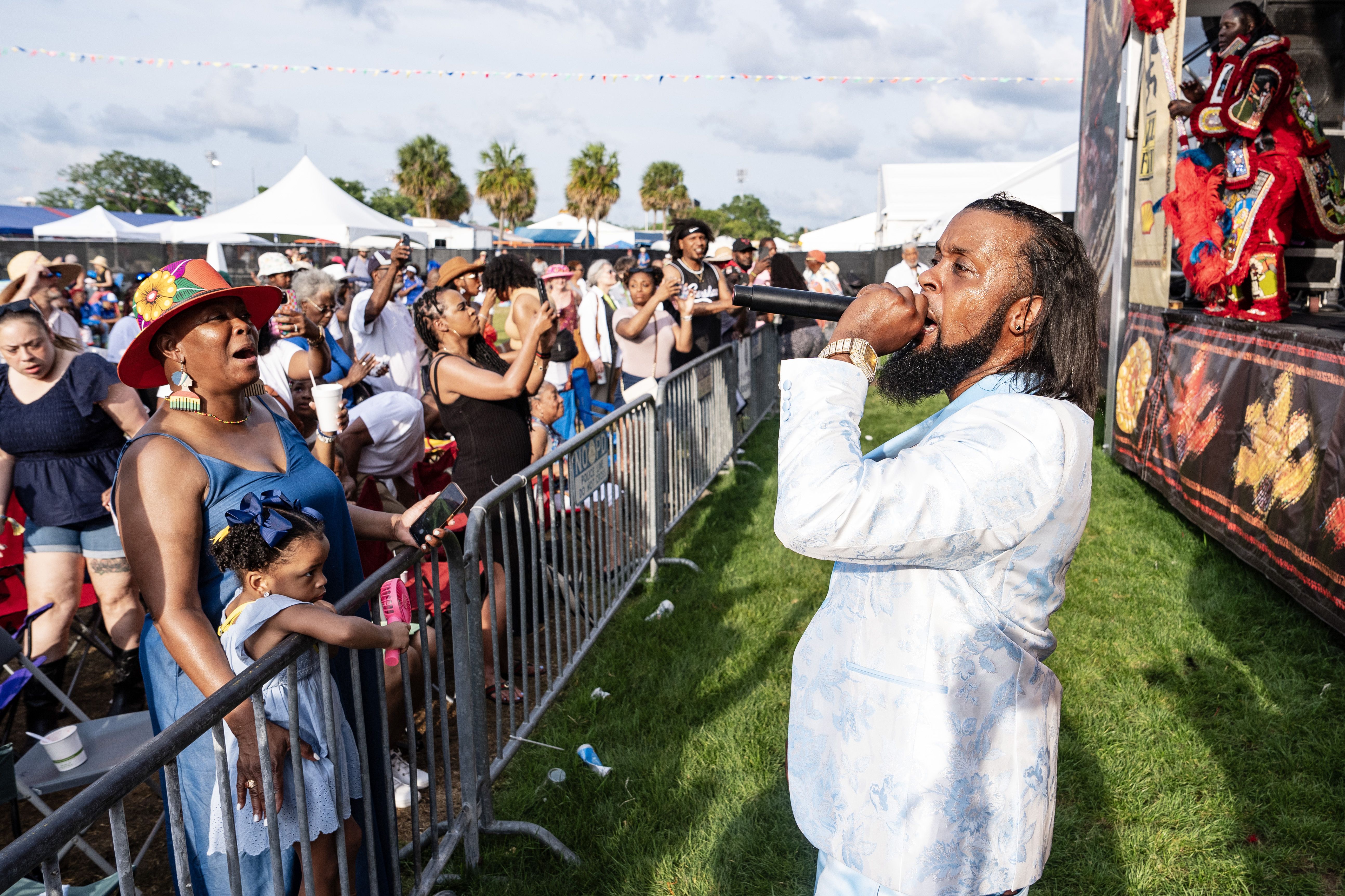 A man sings into a microphone in a green space in front of a stage but behind a barricade, which keeps a big audience at bay.