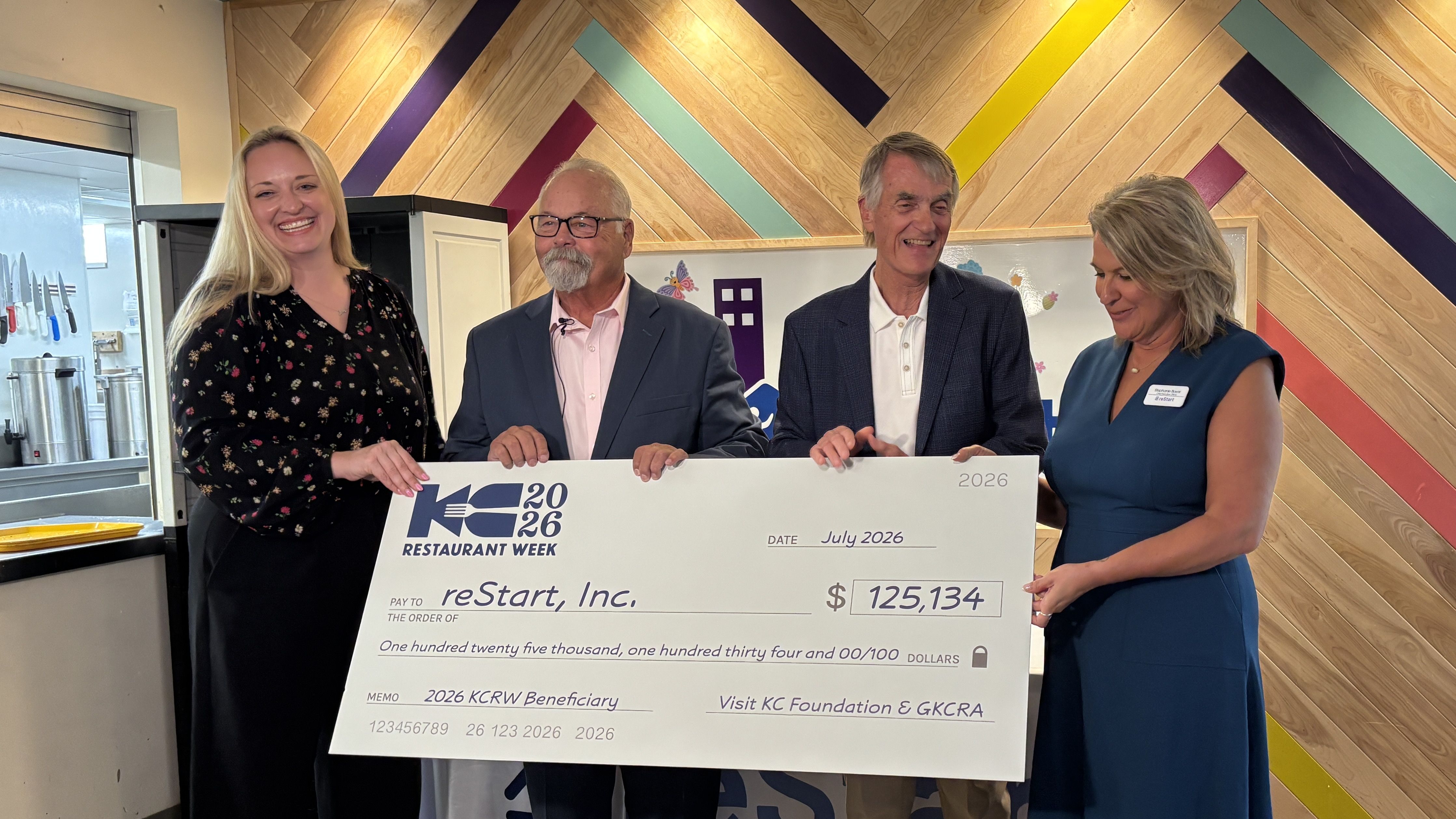 Four people smile while holding a ceremonial check for $125,134 labeled KC Restaurant Week 2026 in a wood-paneled room; left woman wears a floral black blouse, others in business attire.
