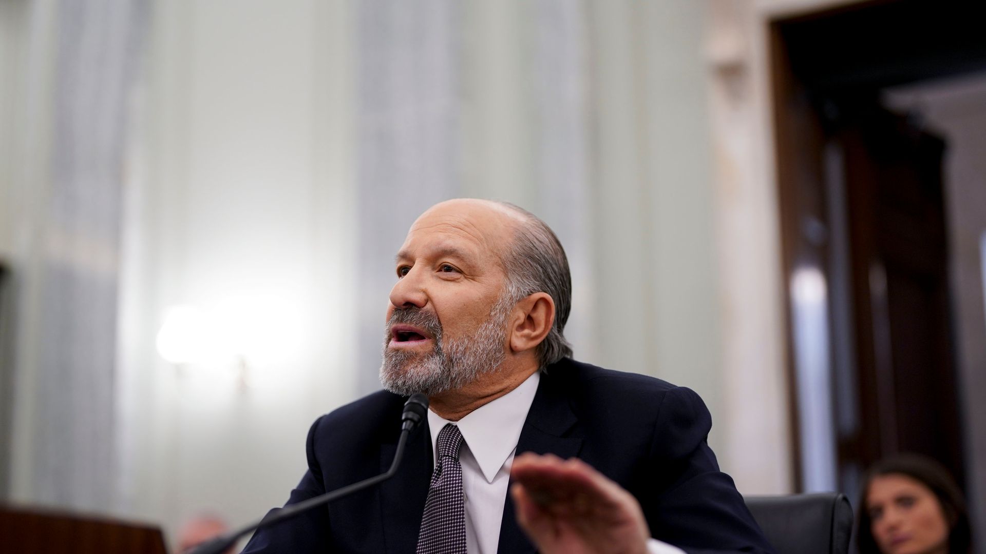 A person speaks in front of a mic wearing a suit and tie