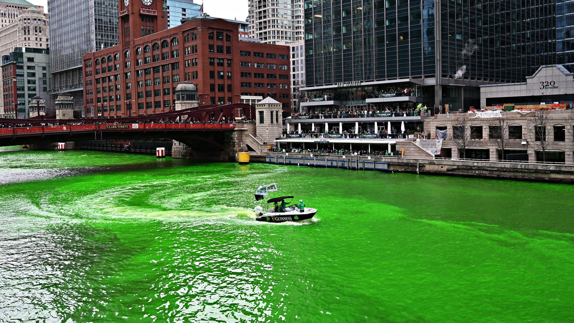 City riverfront scene: neon-green water flows past a red-brick clock-tower building and a red bridge, with modern glass towers and a busy riverside complex; a small Guinness boat sails by.