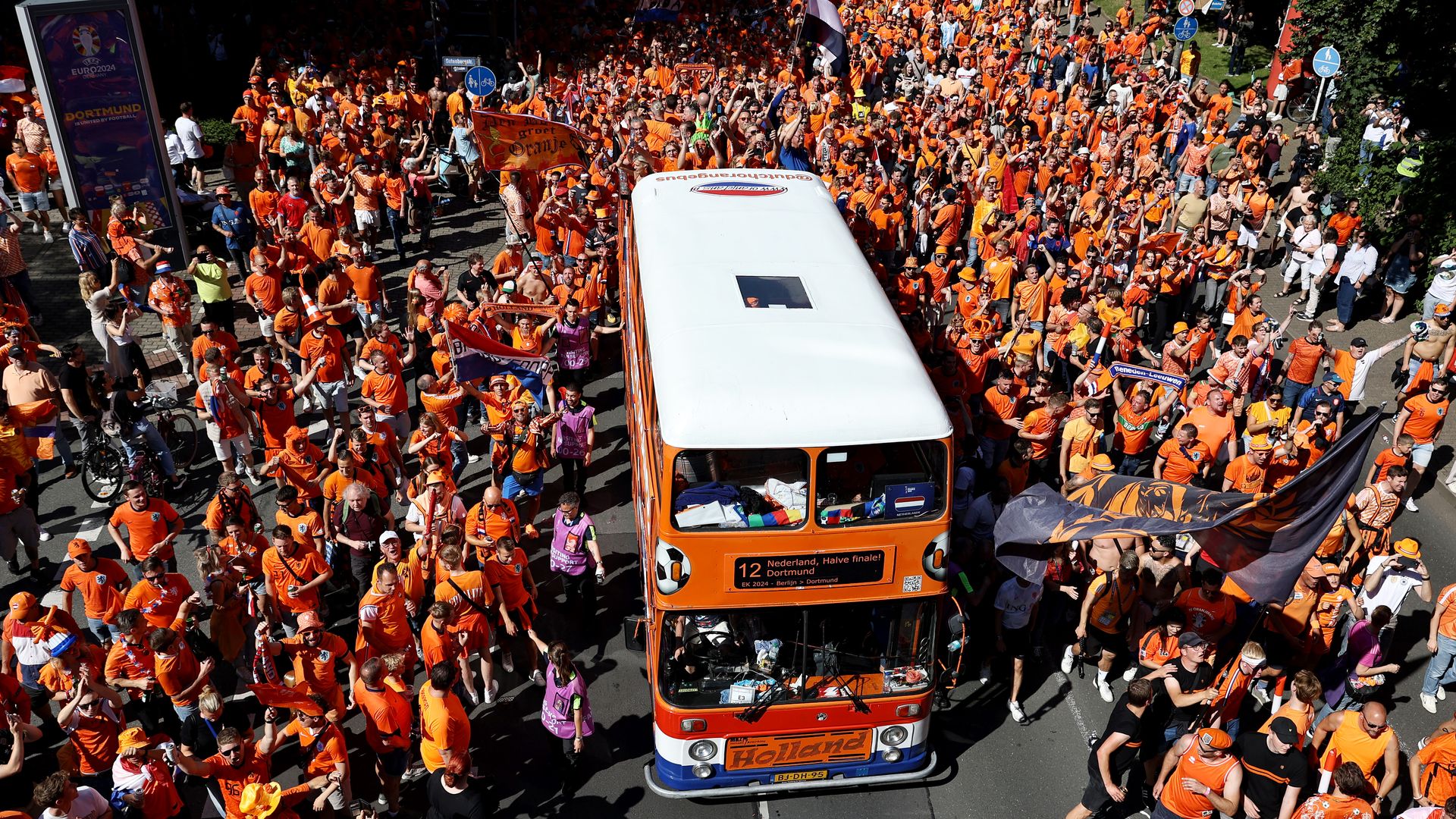 Aerial view of a dense crowd in orange at a street parade, surrounding a white-and-orange bus. Fans wave Dutch flags and banners. 