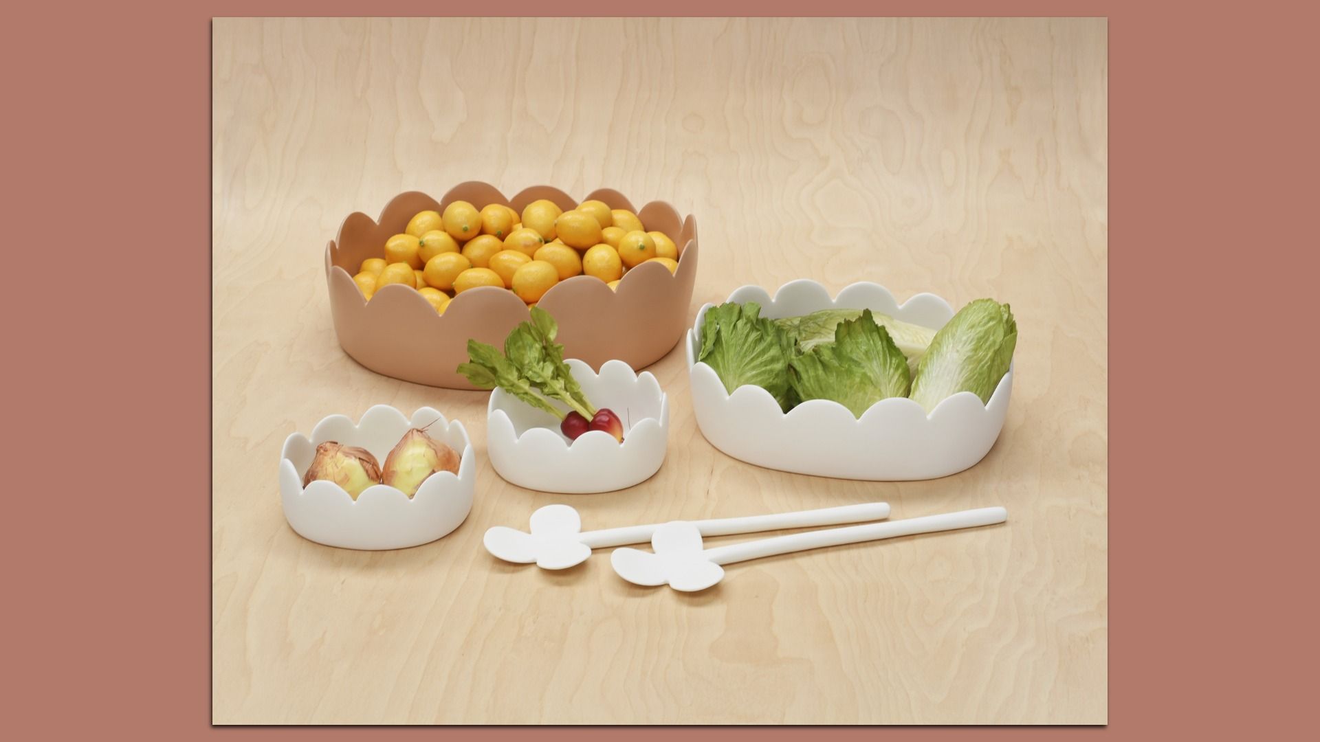 Several scalloped-edge bowls on a light wood surface hold yellow citrus fruits, lettuce leaves and onions; white utensils with flower-shaped ends lie in front.
