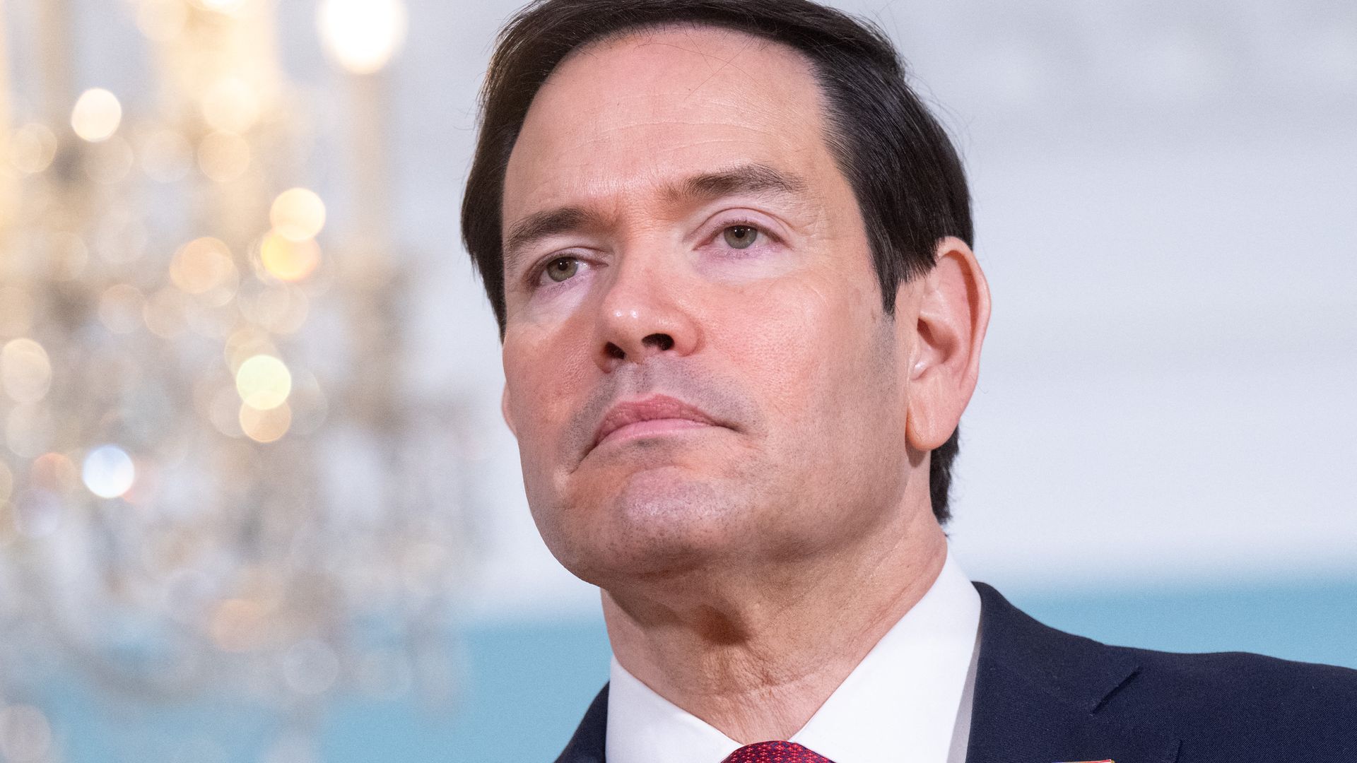 Close-up of Secretary of State Marco Rubio, a middle-aged man in a navy suit, white shirt, red tie, and a small American flag pin on the lapel; neutral expression, soft bokeh background with a chandelier.