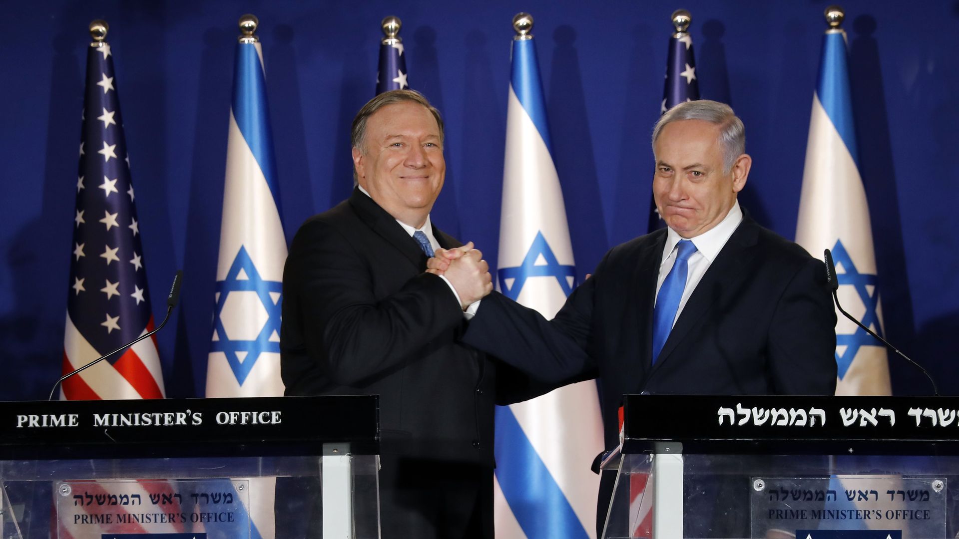 Mike Pompeo and the Israeli prime minister shake hands in front of a row of Israel and American flags, standing behind two podiums. 