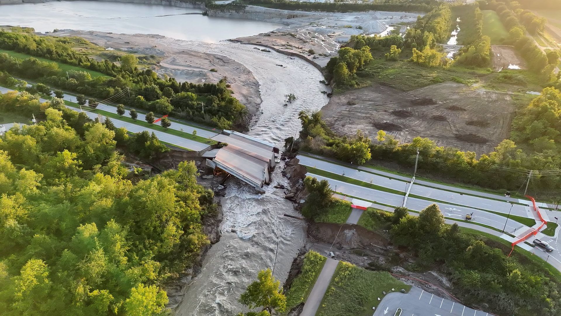 Aerial view of a river cutting through terraced rock with a small bridge, a highway at the bottom, and lush green banks. A distant city skyline sits under a warm sunset glow on the right.