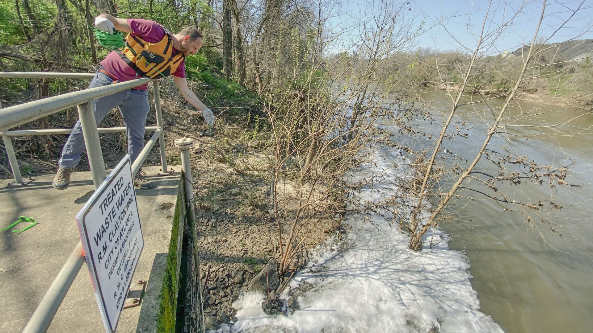 A man wearing an orange vest leans over a railing to conduct water sampling at a treated wastewater discharge site next to a river