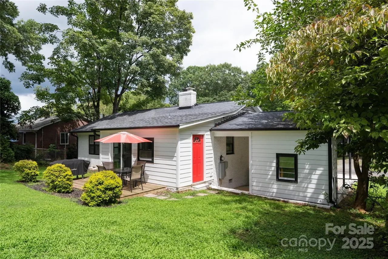 White single-story house with a red door, black roof, small patio with a pink umbrella and table, surrounded by green grass, bushes, and large leafy trees under a partly cloudy sky.