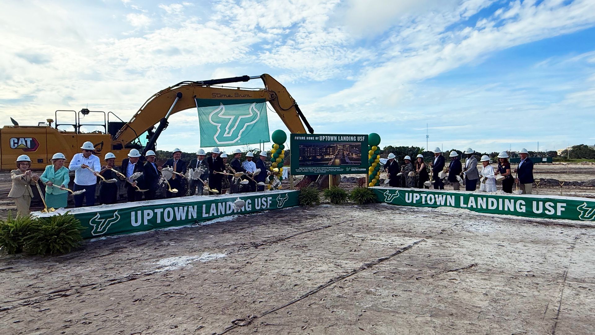 Ribbon-cutting at a construction site with a crowd in hard hats and suits, a yellow excavator, teal USF banner, and a sign reading Future Home of Uptown Landing USF under a blue sky.