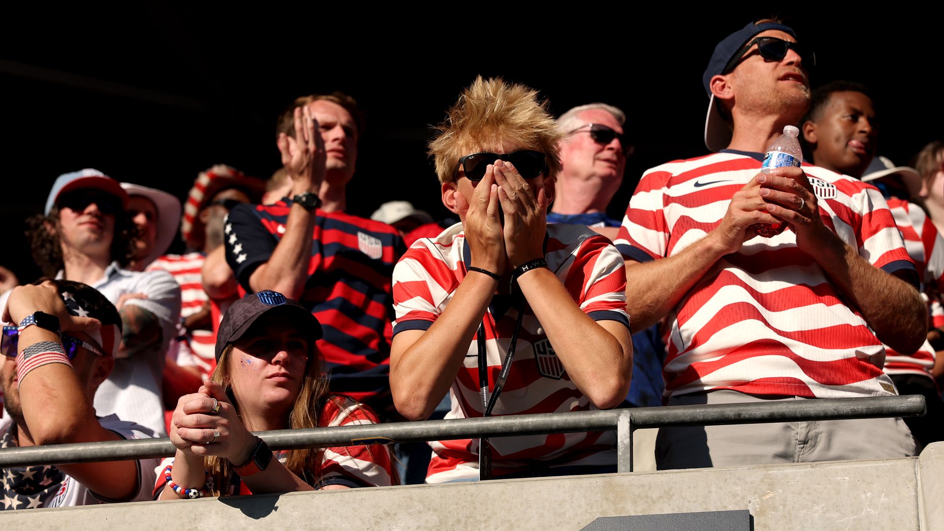 Fans in red-and-white striped U.S. jerseys put their hands to their mouth, clasp their hands and look on with disappointment near the edge of a stadium railing.
