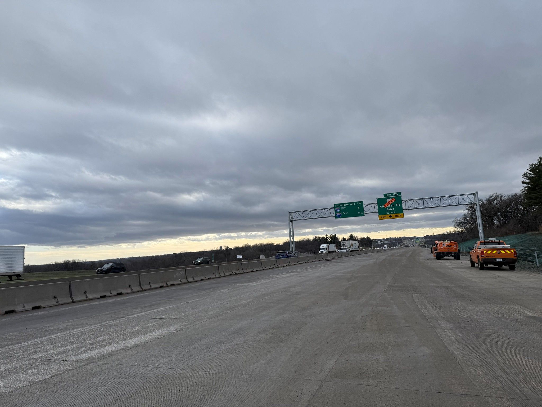 Empty highway with concrete barriers on left, orange construction vehicles on right, under overcast sky. Green highway signs indicate University Ave and exit for Adel Rd.