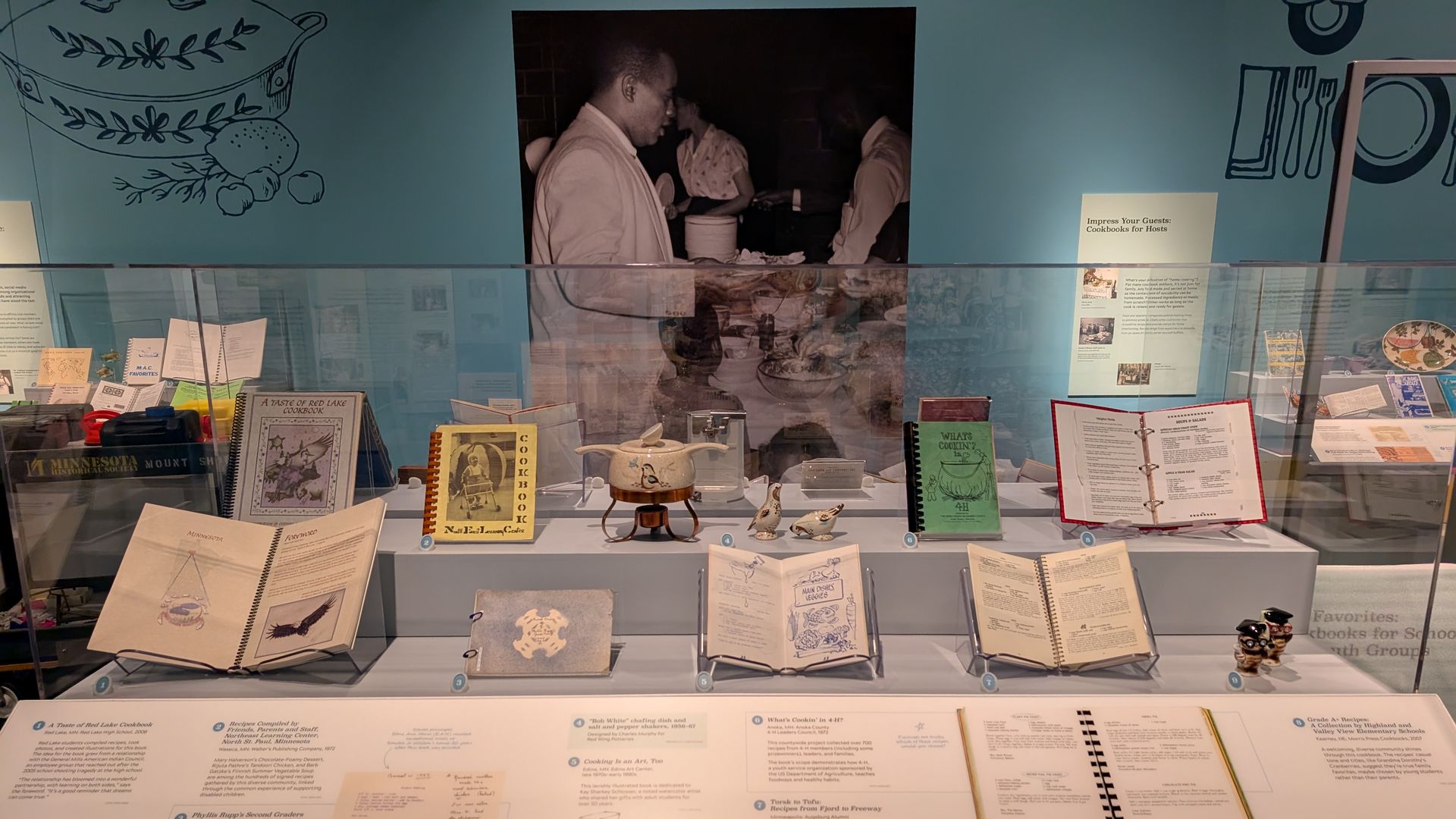 Museum display of vintage cookbooks, ceramic pots, and bird figurines behind glass with informational plaques, set against a blue wall featuring a black-and-white photo of people at a buffet table.