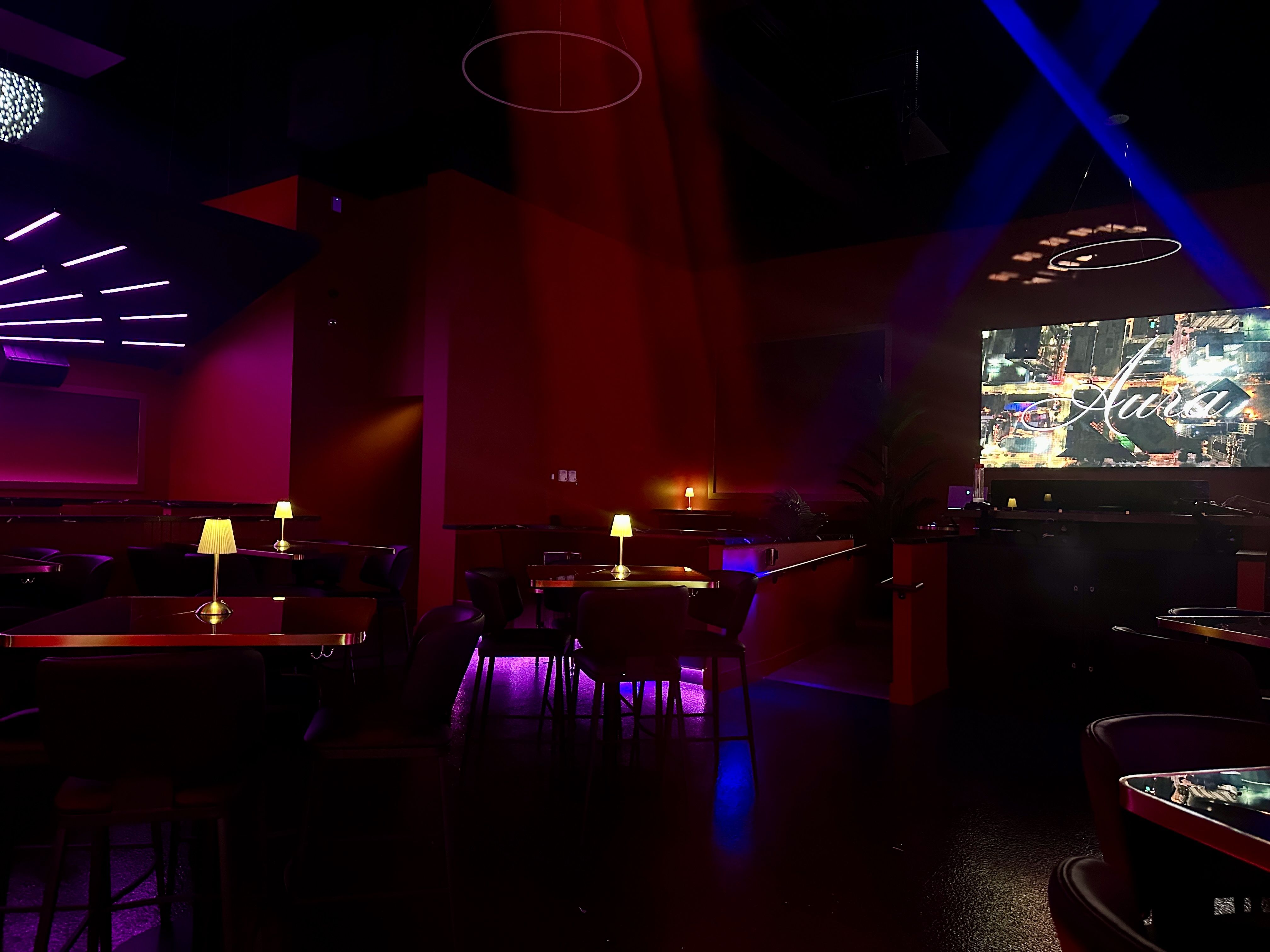 Dimly lit lounge with red walls, tables with small lamps, purple and blue accent lighting, and a large screen displaying the word "Aura" in white script.
