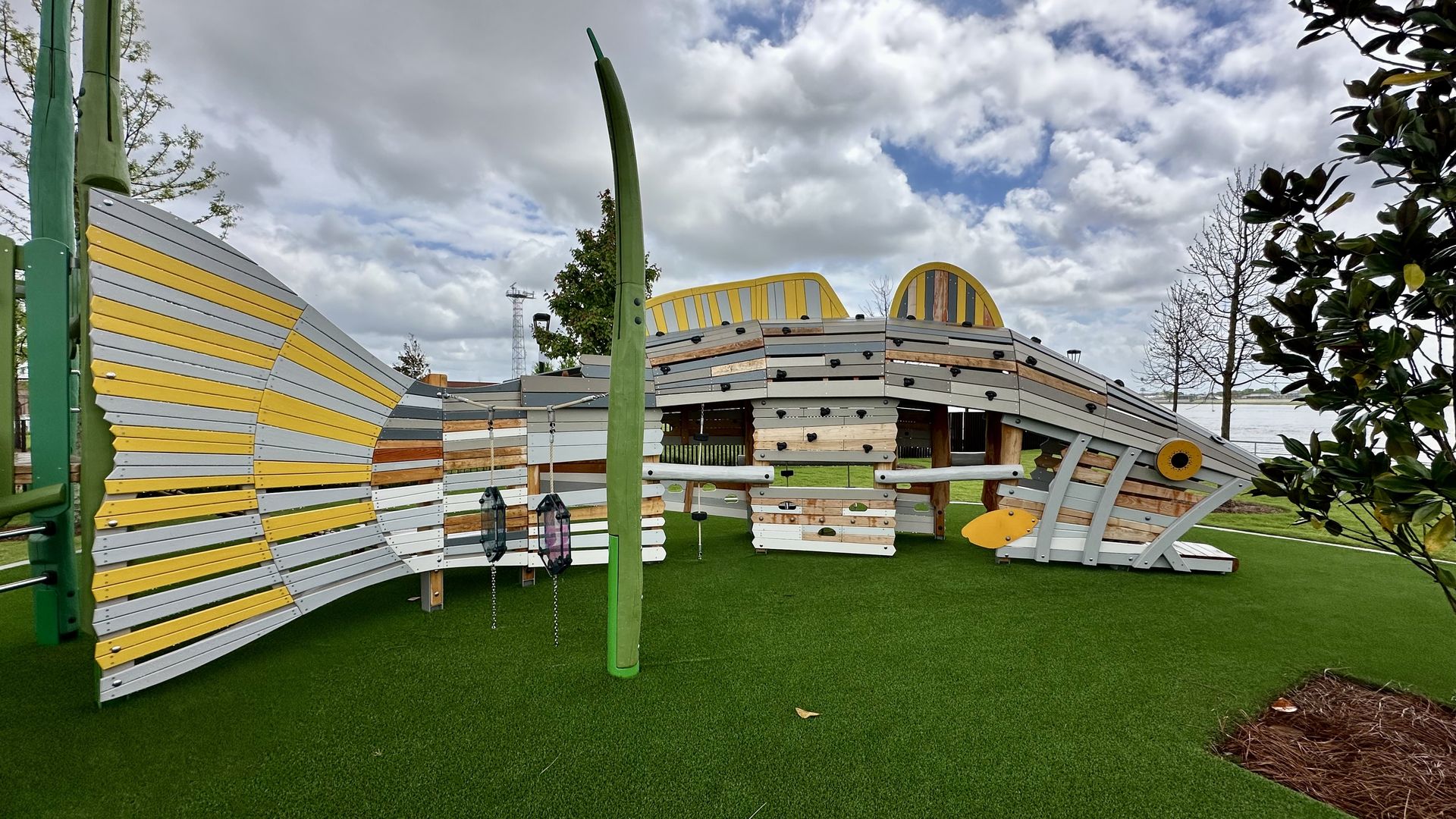 Large playground sculpture built from yellow and gray wooden planks, curved walls, green posts, and swings on artificial turf. A cloudy sky and a body of water visible in the background.
