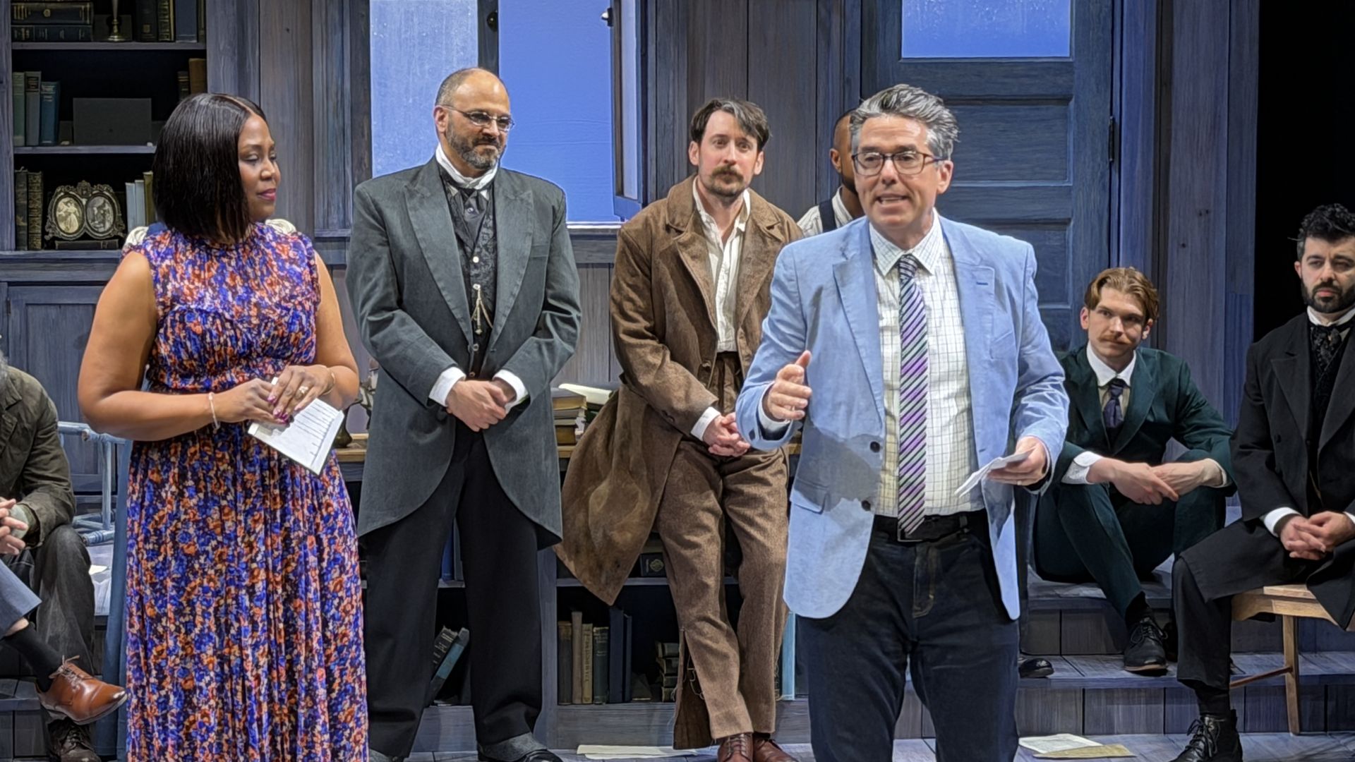 Group of actors in period costumes on a blue-gray stage with bookshelves. A woman in a floral dress stands left; a man in a light blue blazer speaks as others listen.