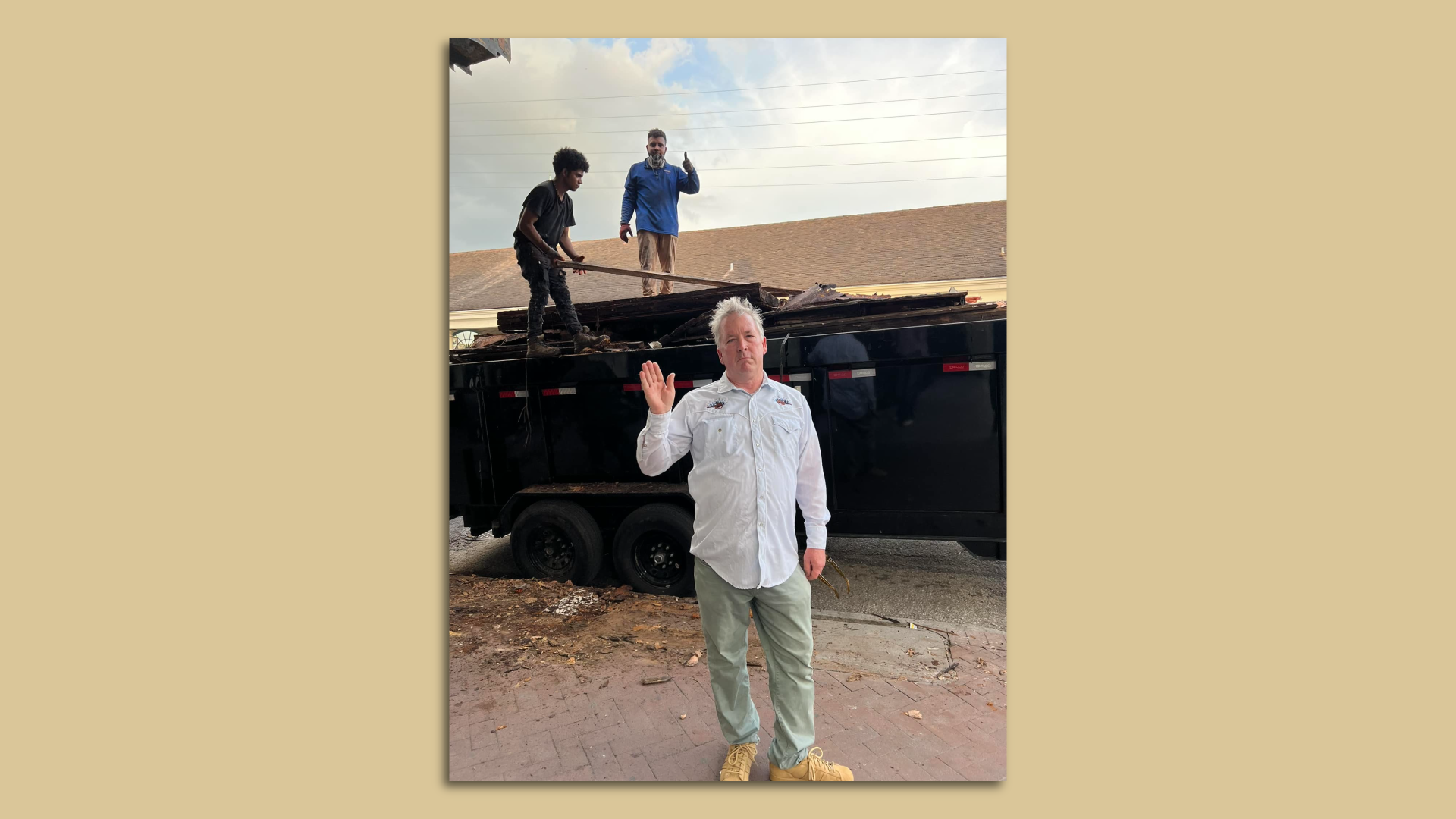 A man stands in front of a trailer bed full of construction debris as construction workers add things to it. The man looks somber and waves to the camera.