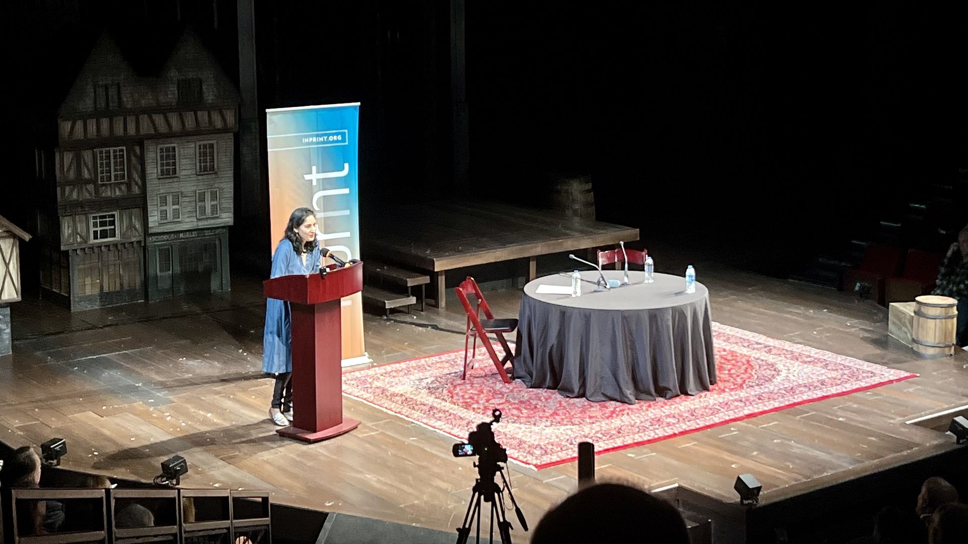 A woman in a blue coat speaks at a red podium on a wooden stage with a patterned red rug. A round table with water bottles and microphones sits nearby; a banner reads "INPRINT."