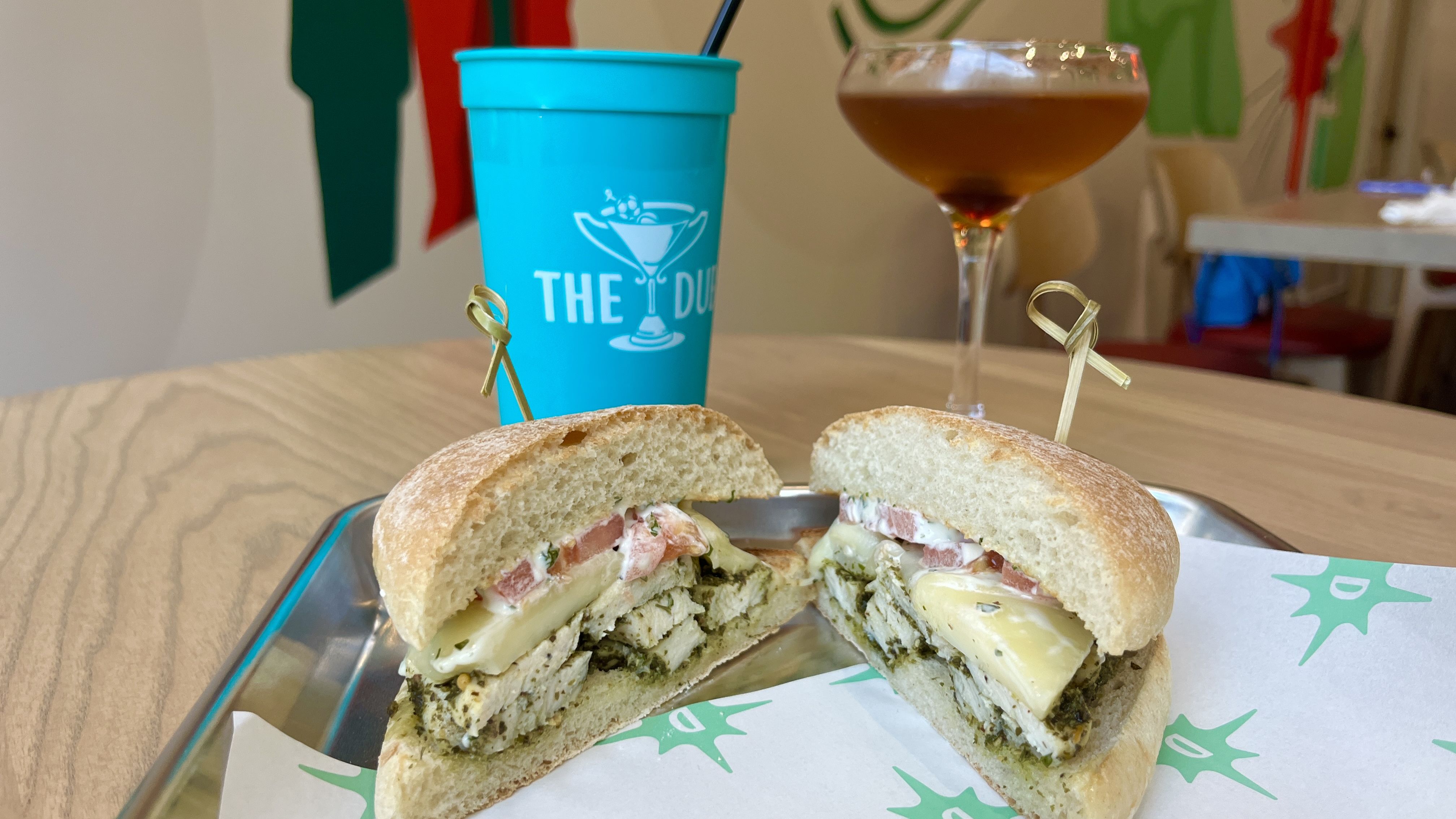 A sandwich cut in half with chicken, cheese, tomato, and sauce on a bun, served on a tray with branded paper. Behind it are a blue cup with a straw and a cocktail glass with a brown drink.