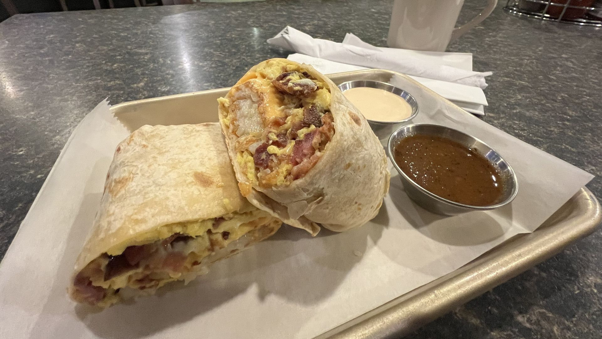 A breakfast burrito on a tray with a small cups of white crema sauce and another small cup of salsa.