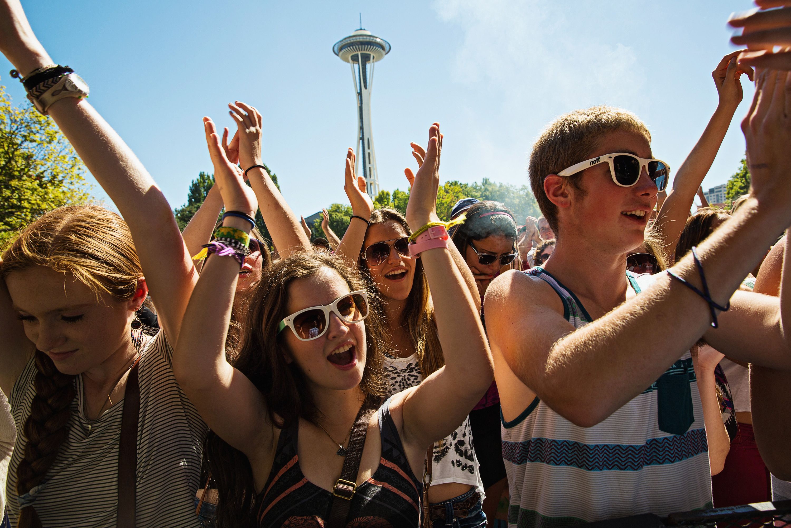 People cheer with their hands up at an outdoor concert with the Space Needle in the background. Several of the concert goers are wearing sunglasses and tank tops.
