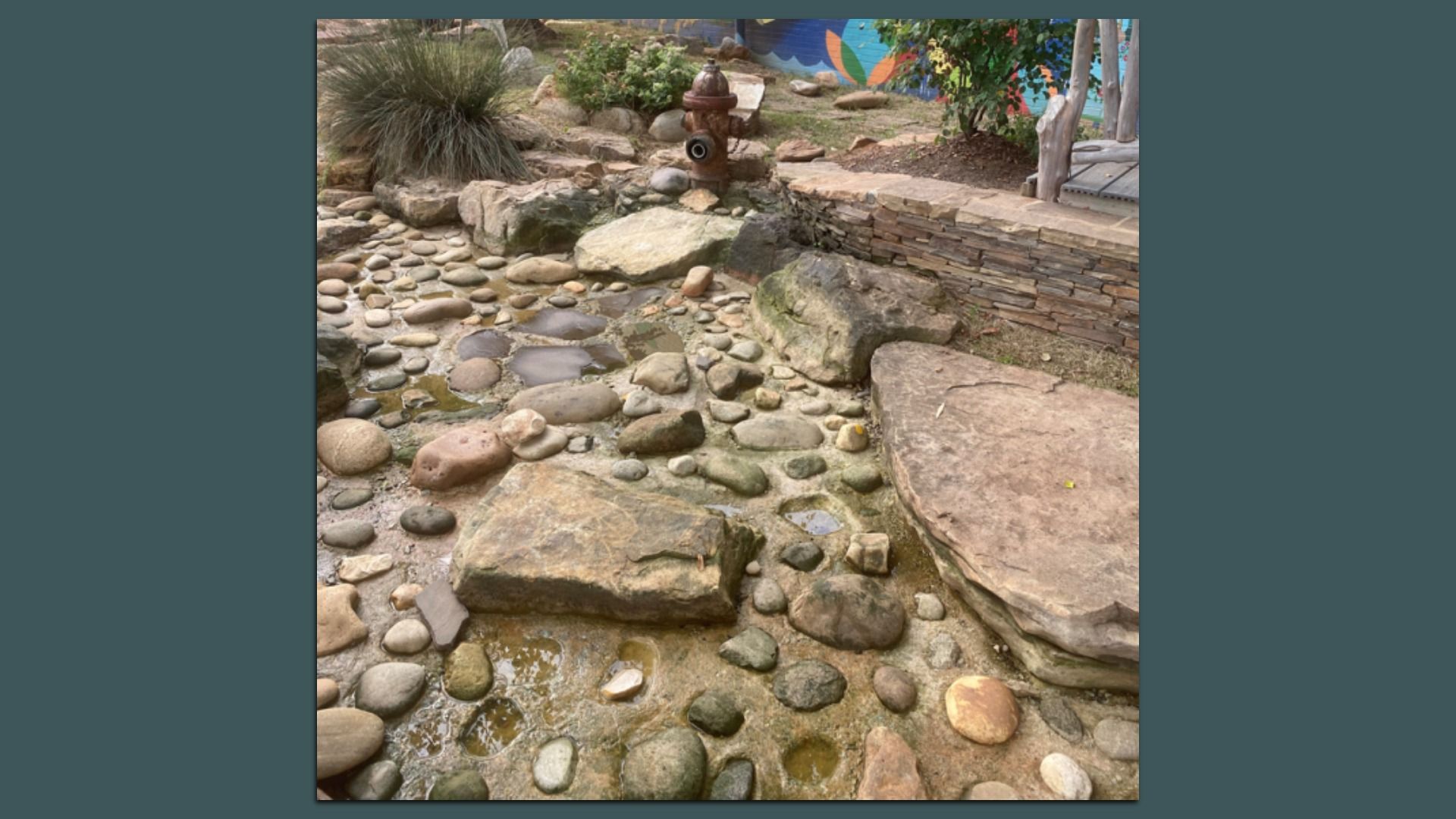 Rocky garden with a dry creek bed of smooth pebbles, large stones and a stacked stone wall on the right. A water hydrant centerpiece sits among greenery, with a colorful mural in the background.