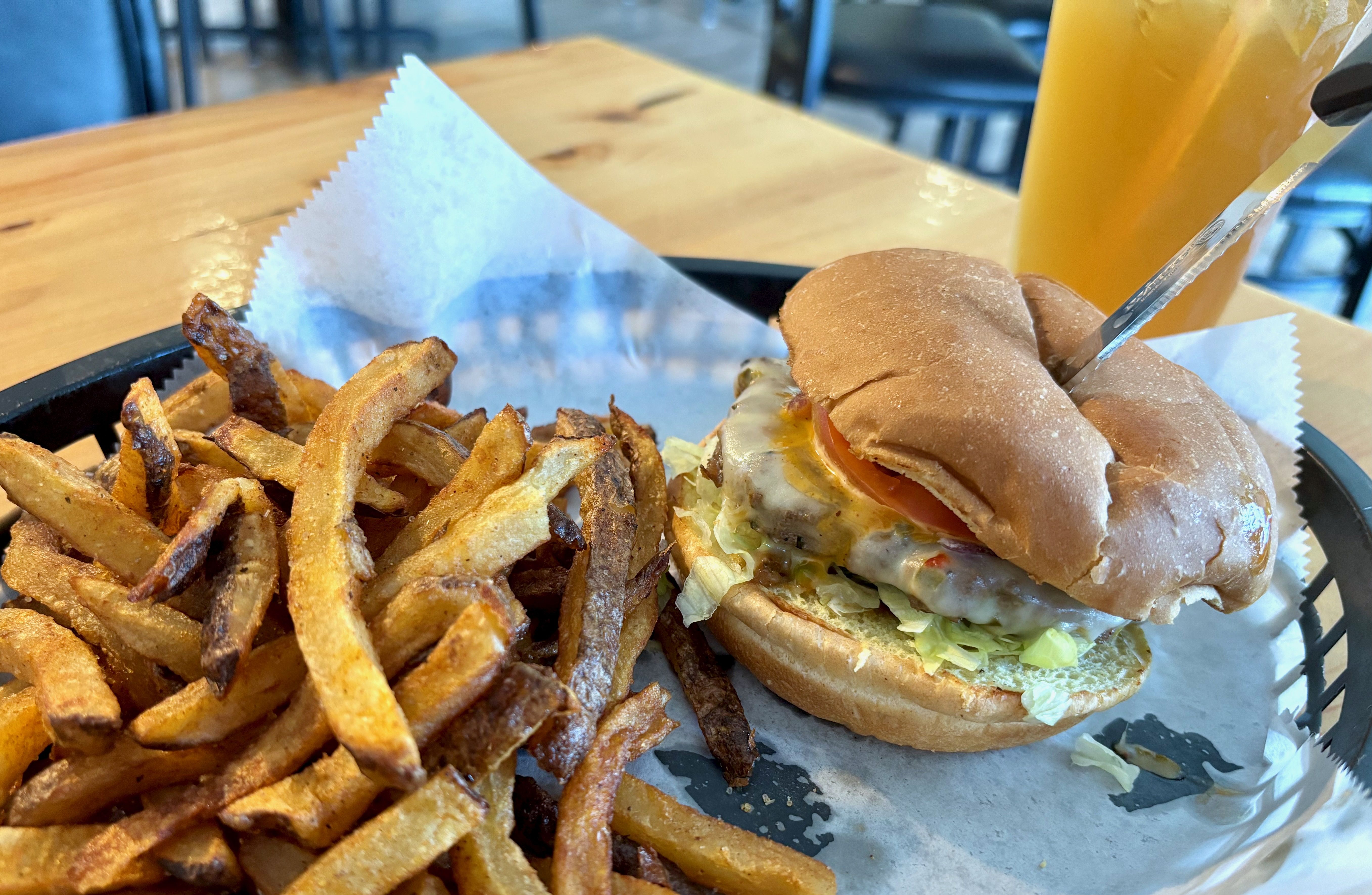Cheeseburger with lettuce and tomato on a bun, served with crispy golden fries on a black tray with white paper, with an iced orange drink in the background on a wooden table.