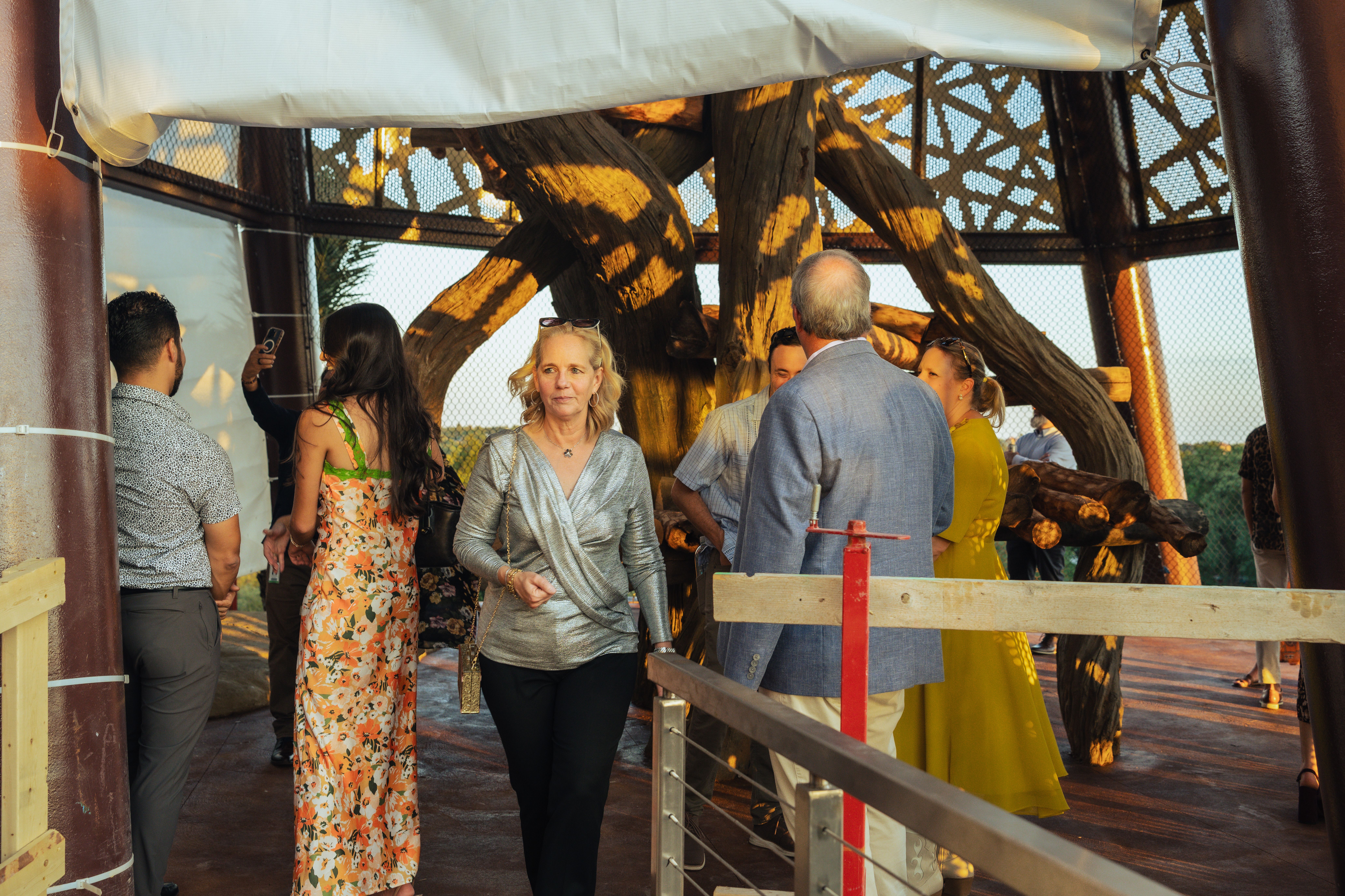 Group of people dressed in semi-formal attire inside a structure with large wooden beams and mesh walls, bathed in warm late afternoon sunlight.