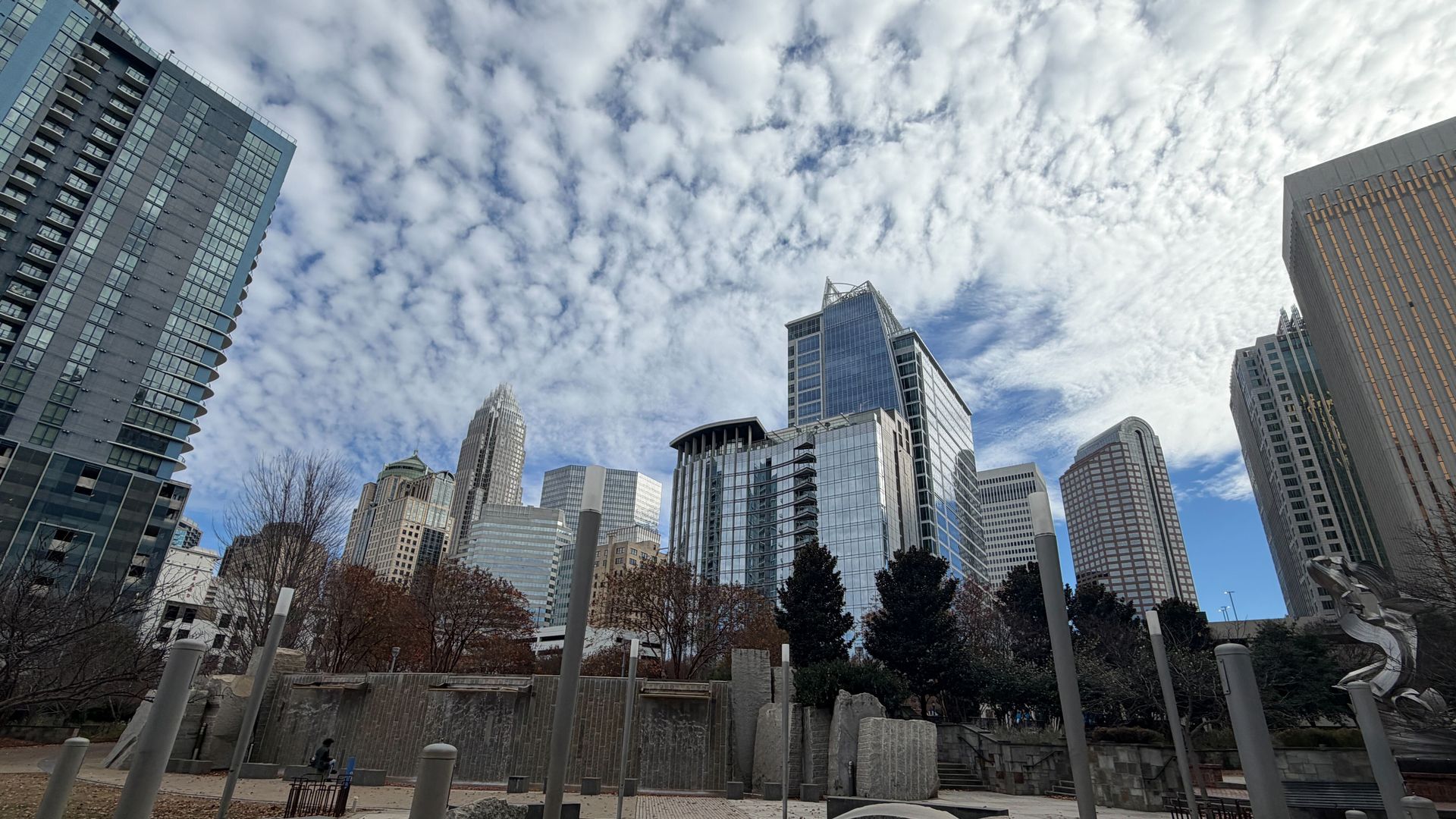 Charlotte skyline. Wide cityscape of modern glass skyscrapers under a blue, partly cloudy sky; foreground shows a paved walkway, green grass, large rocks, and numerous tall gray posts in a plaza.