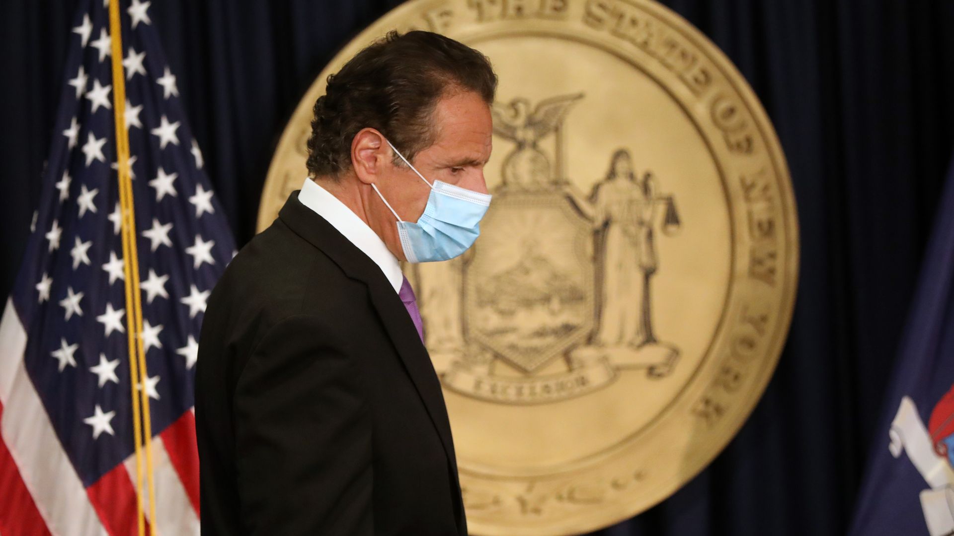 Photo of a masked Andrew Cuomo looking down, an American flag and seal behind him