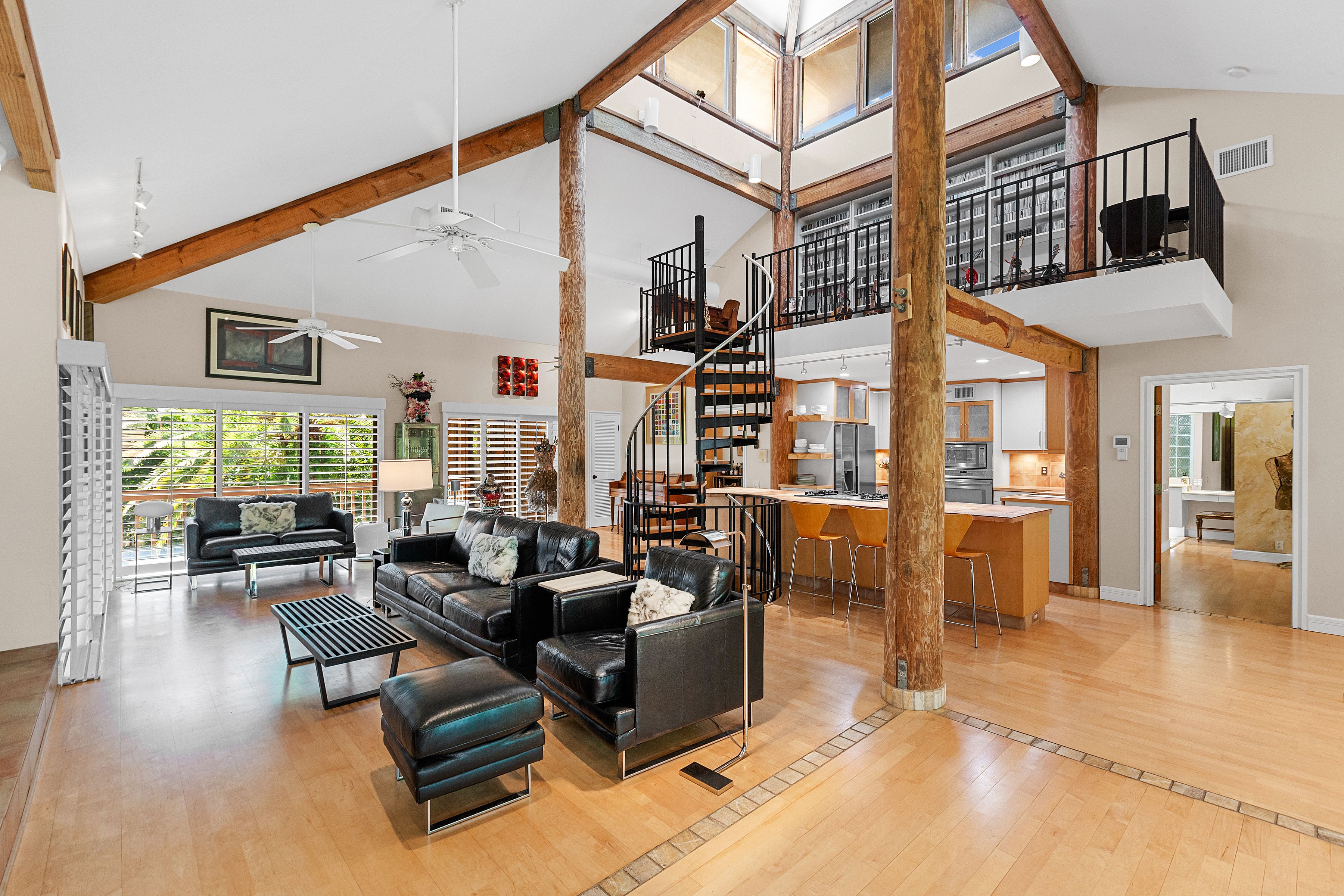 A living room and kitchen decorated with mid-century-style furniture and exposed wood beams. A spiral staircase from the kitchen leads up to a library with built-in bookshelves.