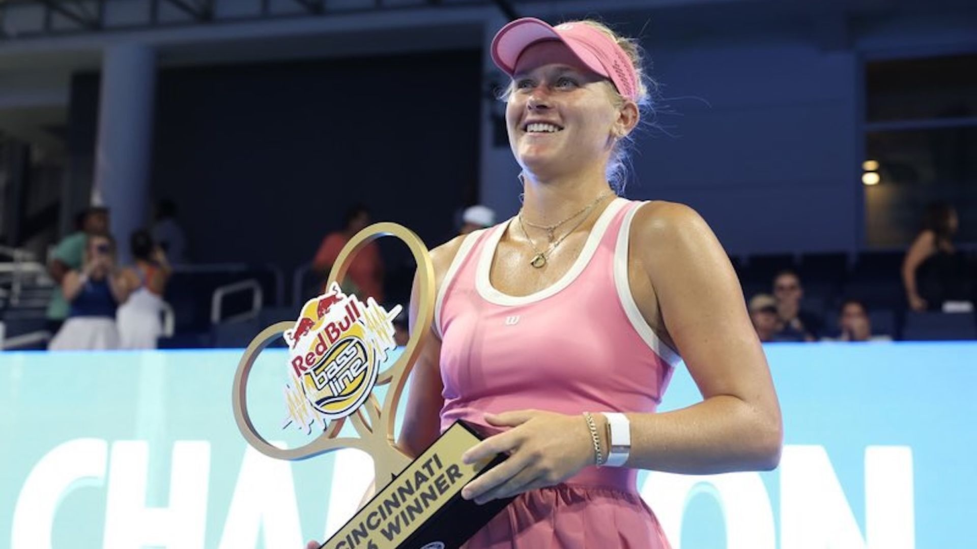 Smiling female athlete in a pink tennis outfit and visor holds a trophy with a Red Bull logo that reads "Cincinnati 2026 Winner", against a blue arena backdrop.