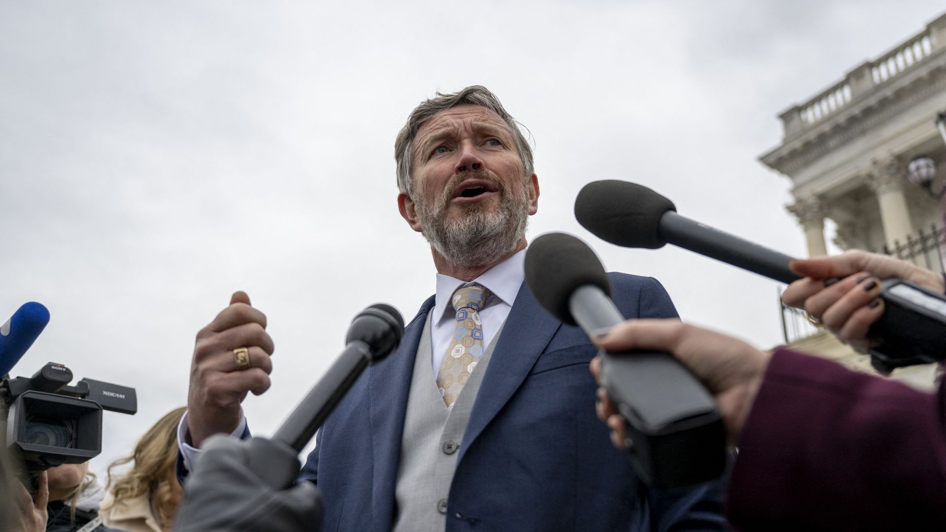 Man in a blue suit and patterned tie speaking passionately to reporters holding microphones with a government building in the background under a cloudy sky.