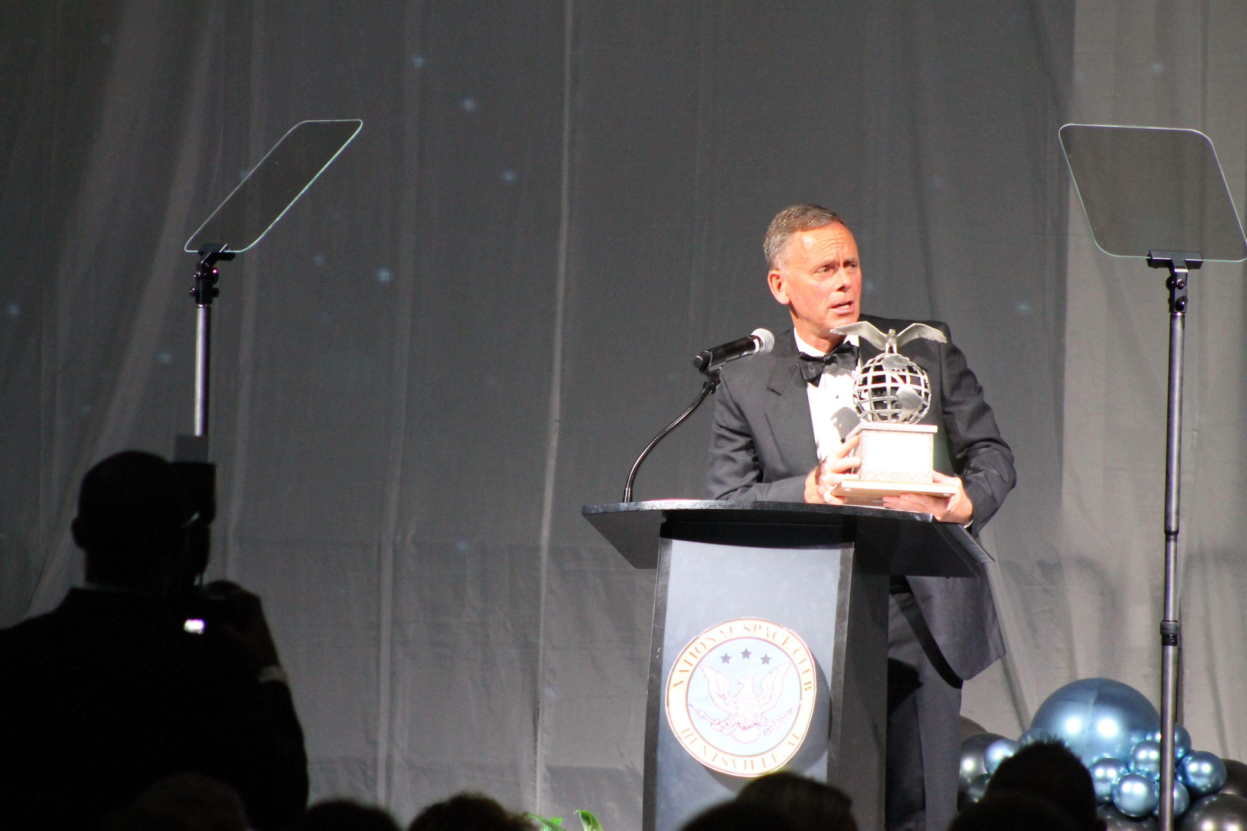 A man in a black tuxedo holding a globe-shaped award with wings stands behind a podium labeled "National Space Club Huntsville." Two teleprompters and blue balloons are in the background.