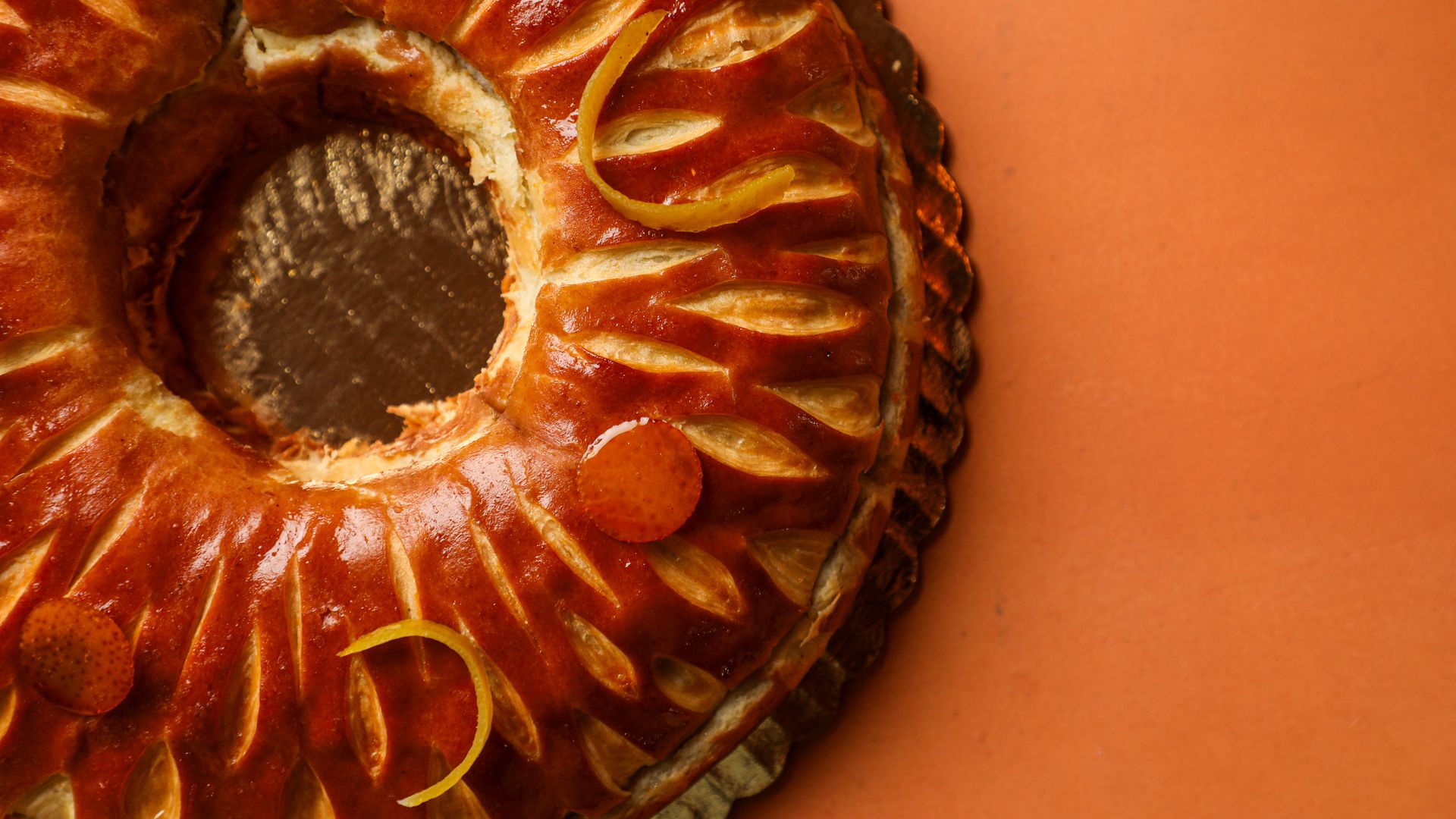 Close-up of a shiny, golden-brown circular bread with decorative cuts and candied orange peel on top, placed on a textured orange surface.