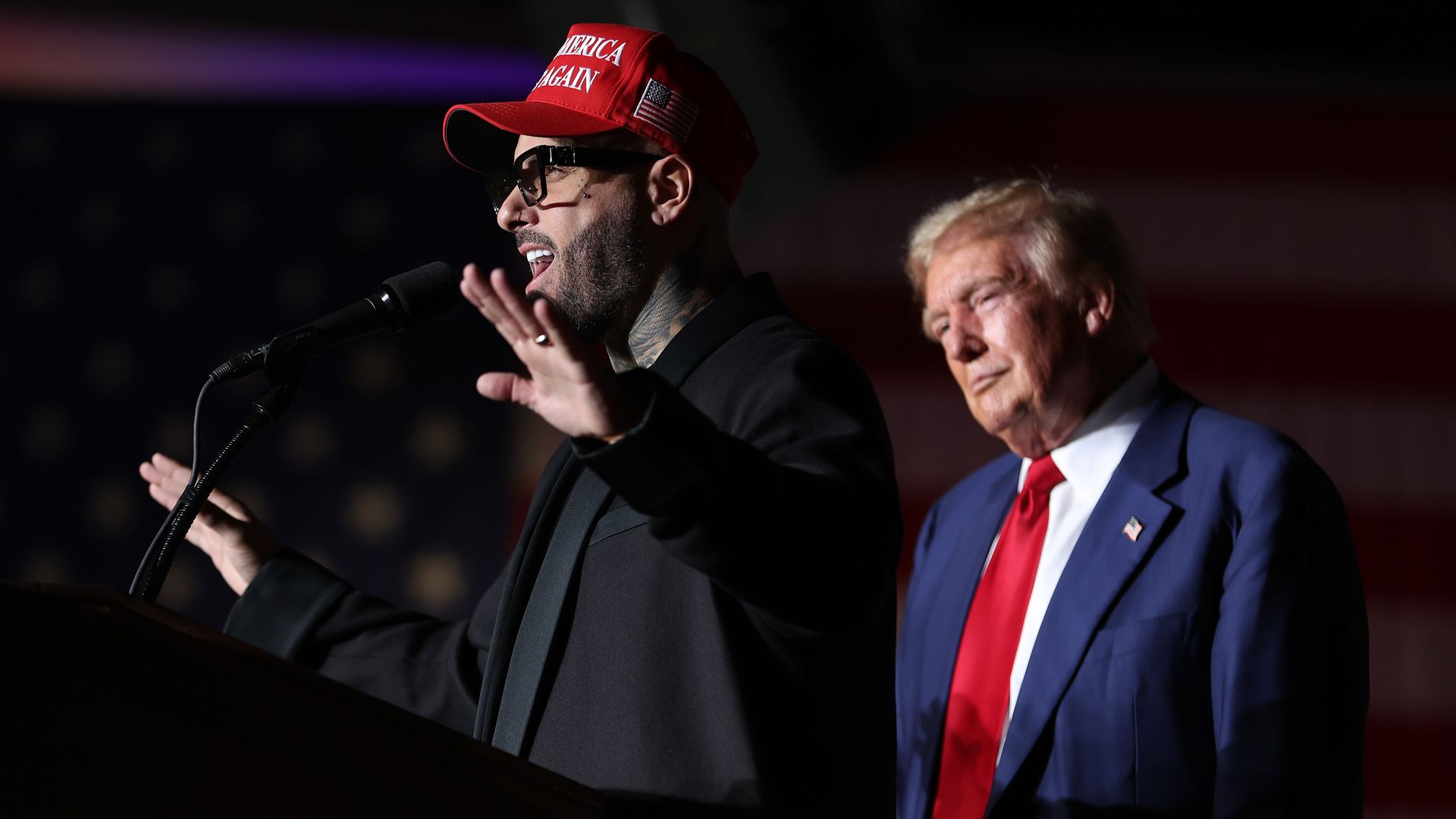 Nicky Jam, wearing a Make America Great Again hat, speaks in front of a podium while former President Trump stands behind him. 