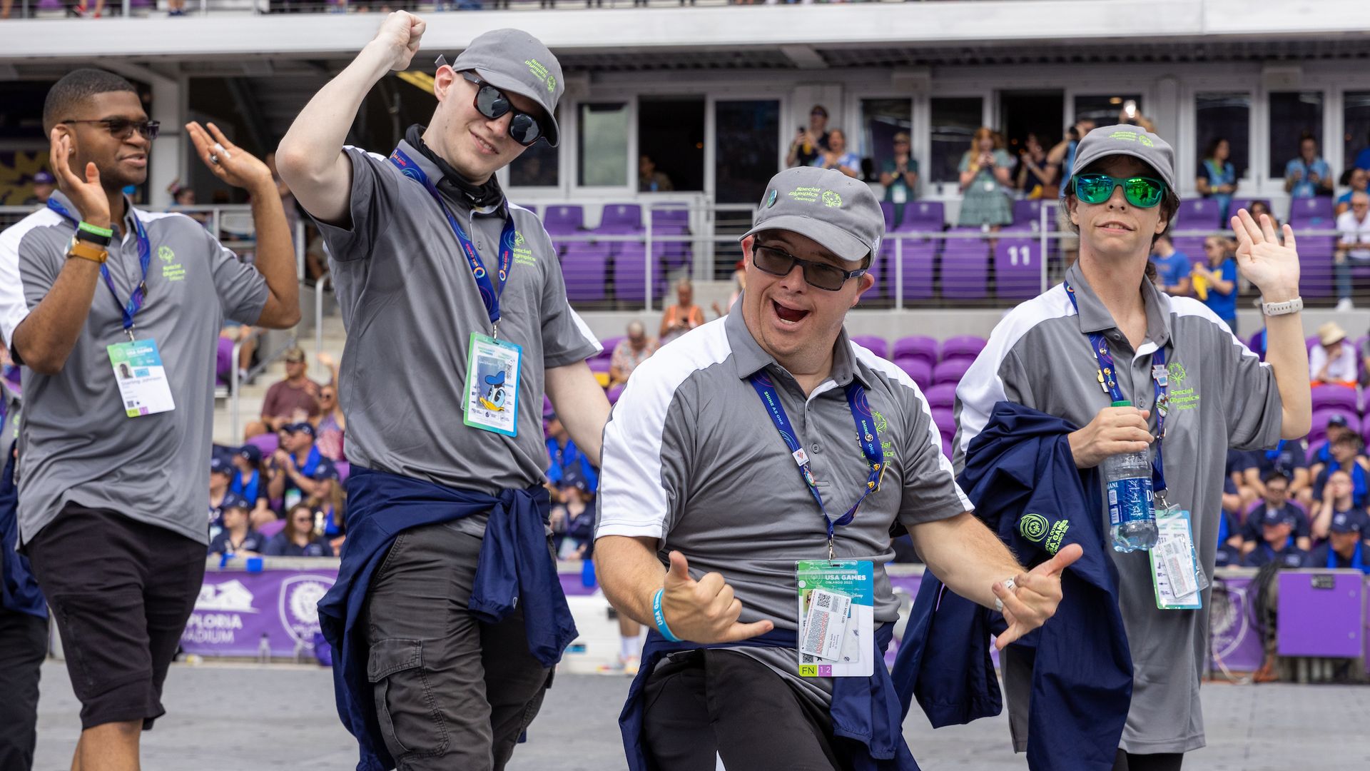 Four volunteers in gray uniforms and caps cheer, wave, and pose at a stadium with purple seating; they wear lanyards and sunglasses as spectators watch.
