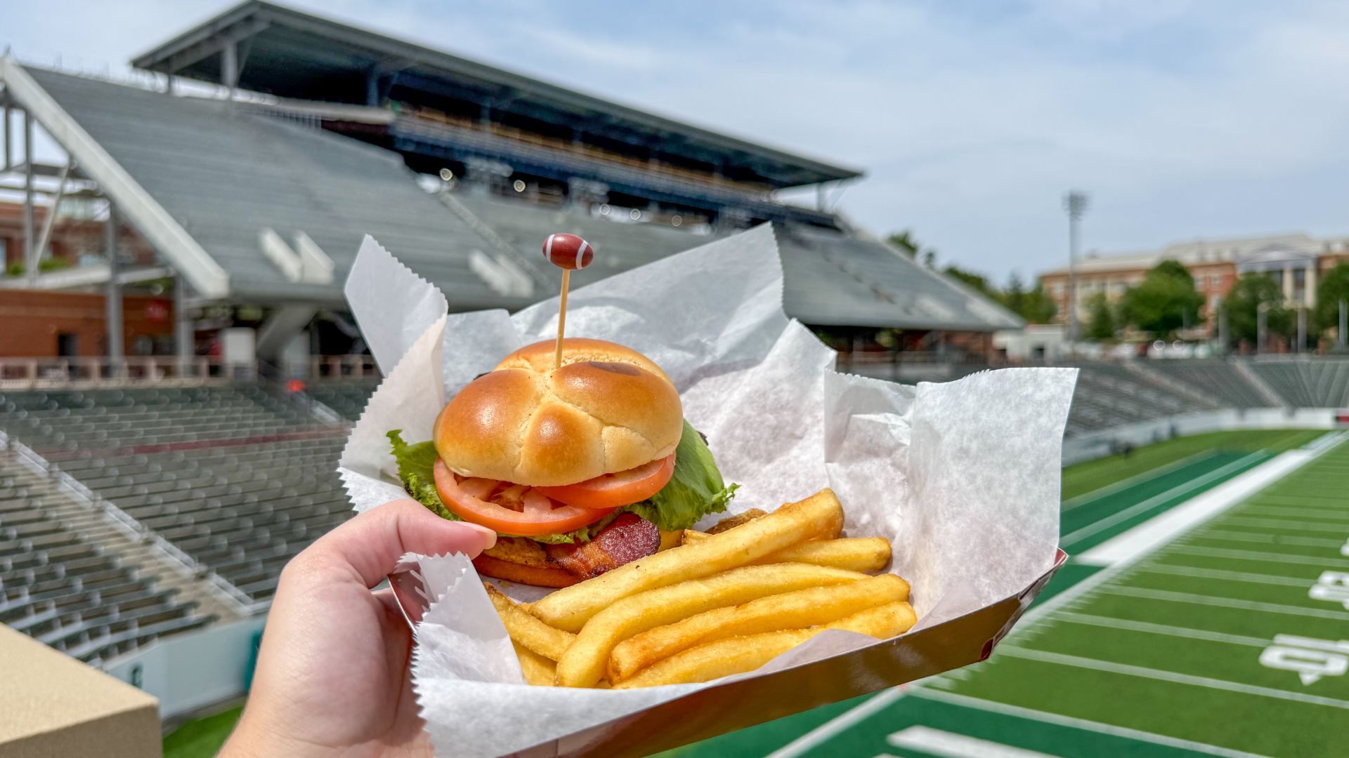 Hand holding a tray with a hamburger (bun, lettuce, tomato, bacon) and french fries, in front of stadium seating and a green football field.