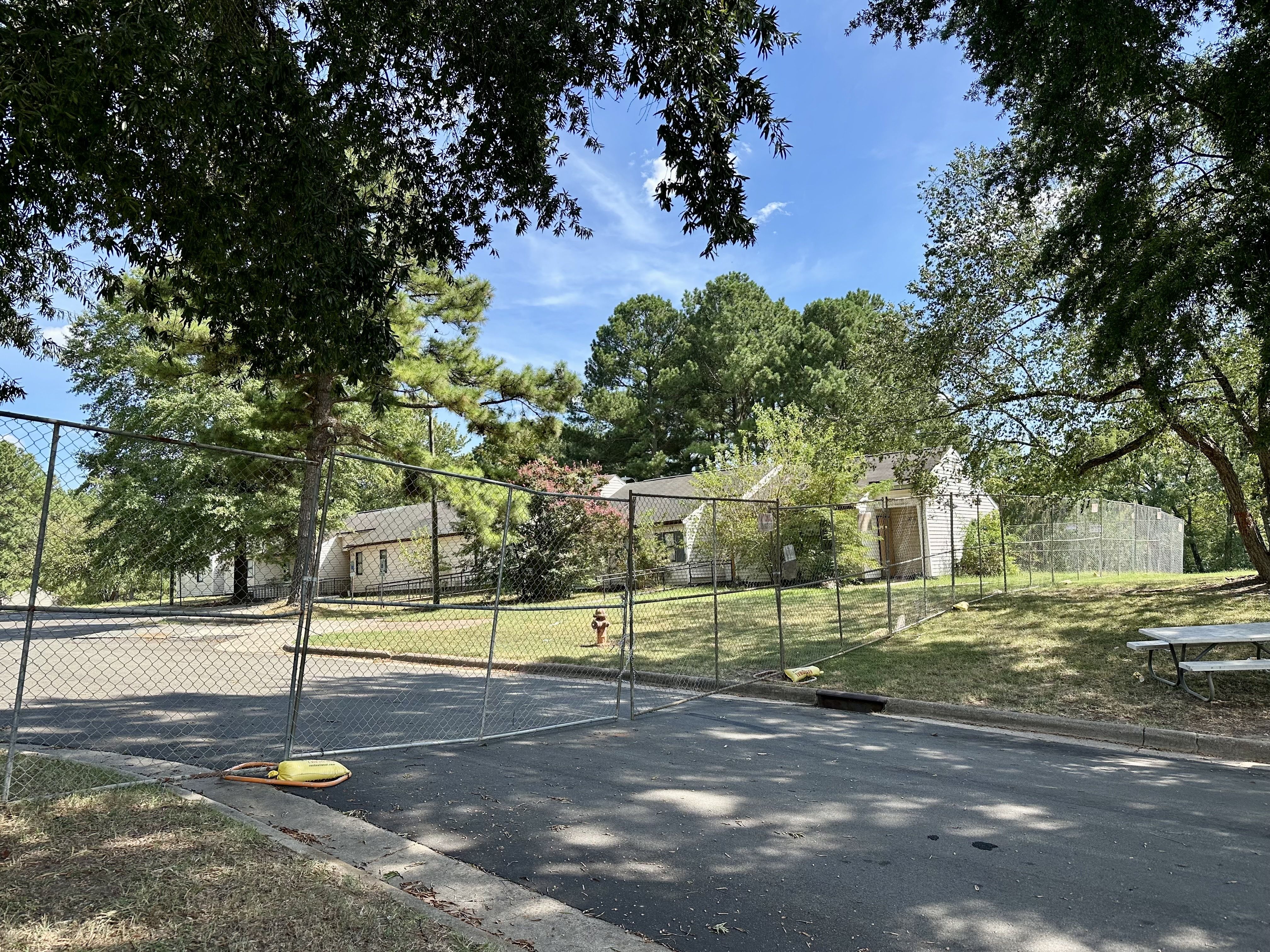 Wide view of a chain-link fenced park area beneath a dense tree canopy with a bright blue sky; houses are in the background, a picnic table on the right, and a yellow object on the pavement near the fence.