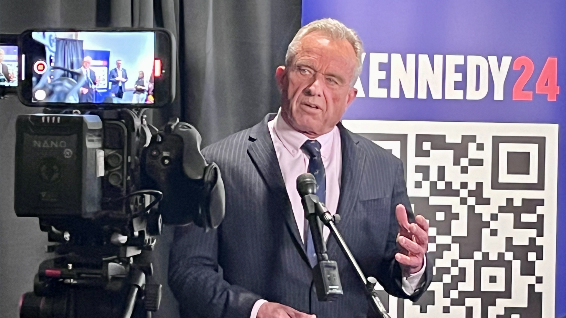 A political candidate speaking at a press conference, with white hair and a grey suit. He's gesuring with one hand as he looks toward a camera in the foreground.