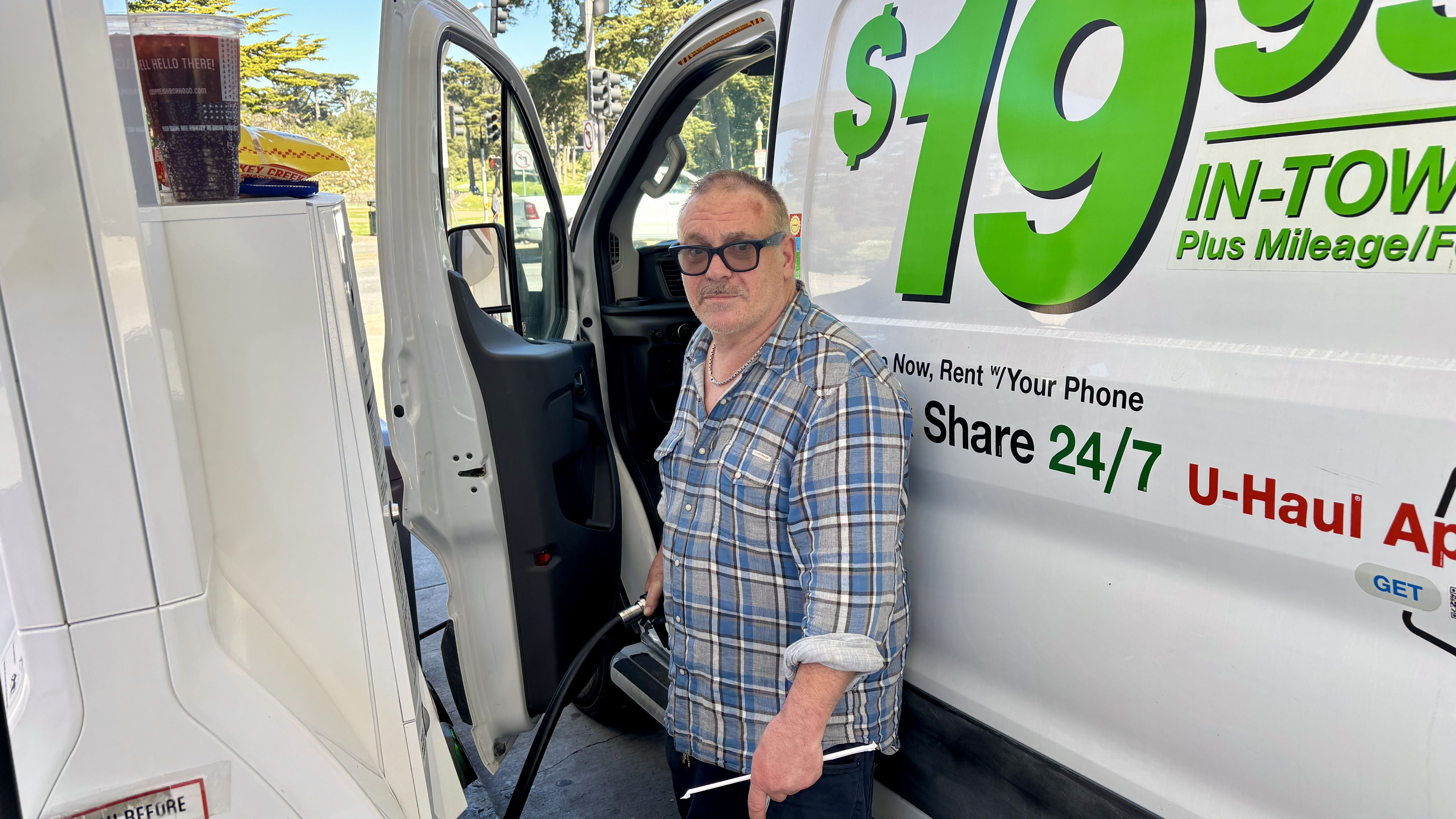 Man in a blue plaid shirt and sunglasses stands by the open door of a white van with large green numbers, promoting "Share 24/7 U-Haul App" while holding a hose on a sunny street.