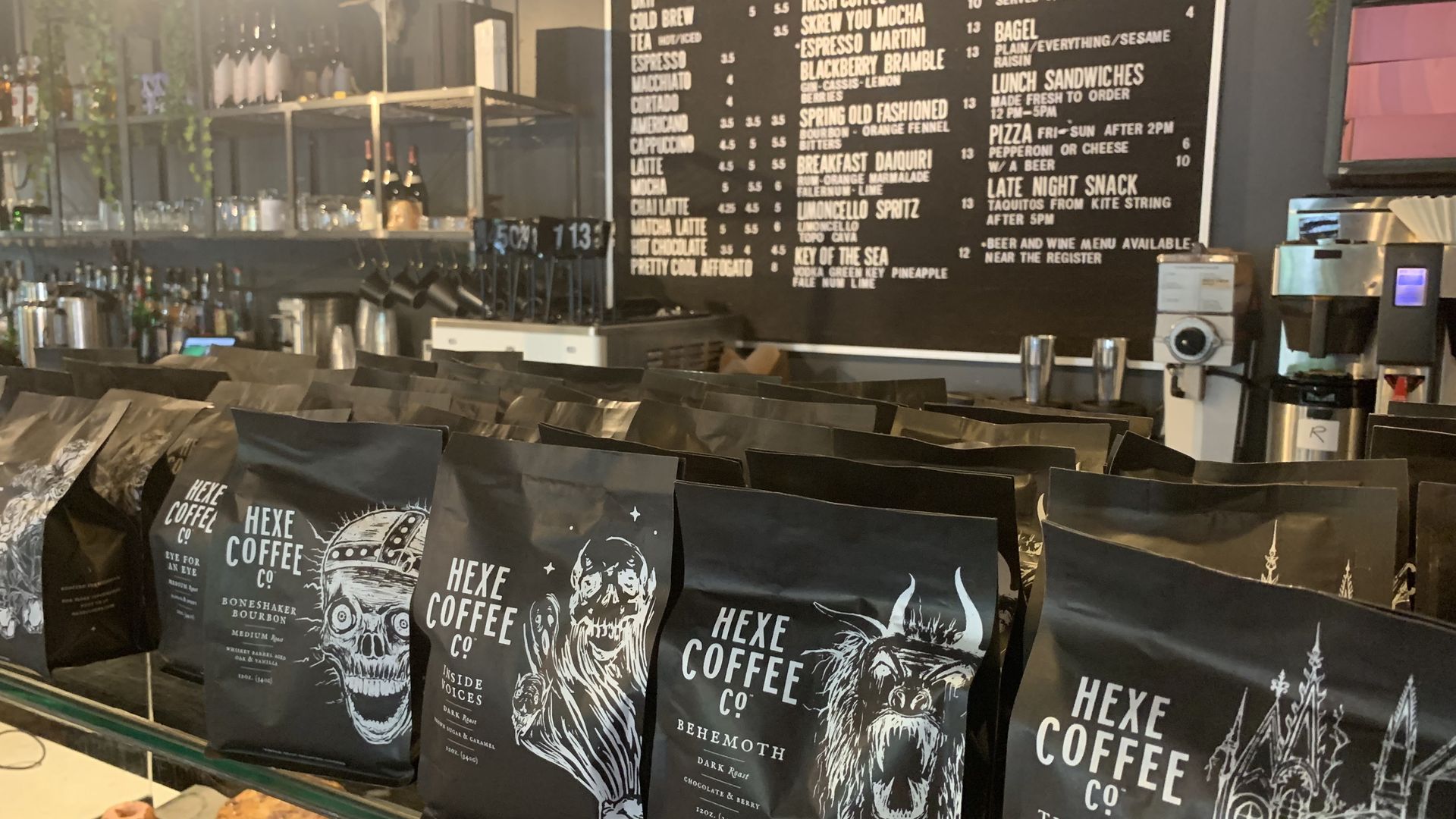 Interior of a cafe with a large black menu board listing coffee and food; foreground features Hexe Coffee Co. bags with skull art and a pastry display; warm light and hanging plants.
