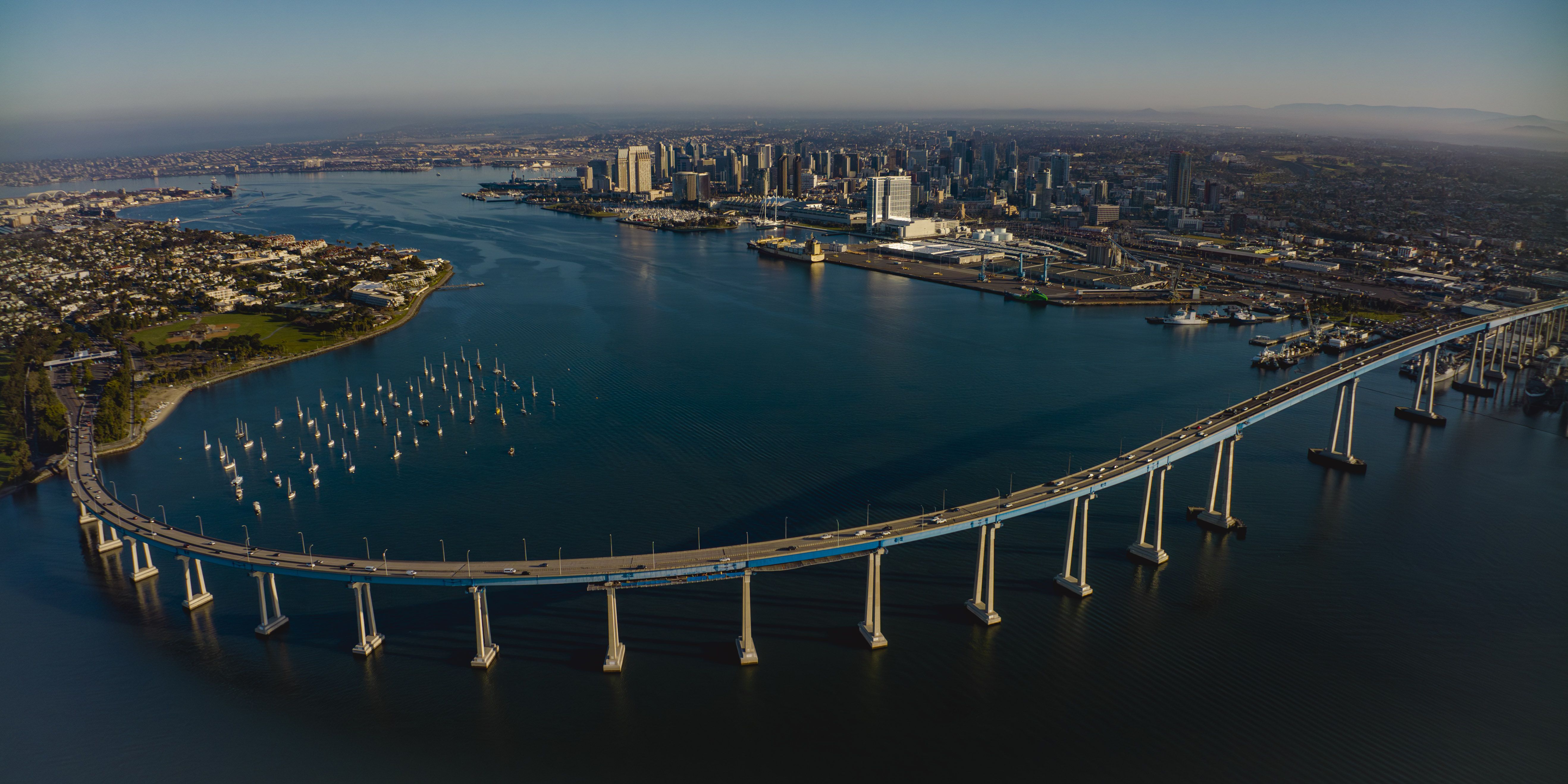 Aerial view of a long curved bridge over blue water, with many sailboats anchored near a green area and a city skyline with tall buildings in the background under a clear sky.