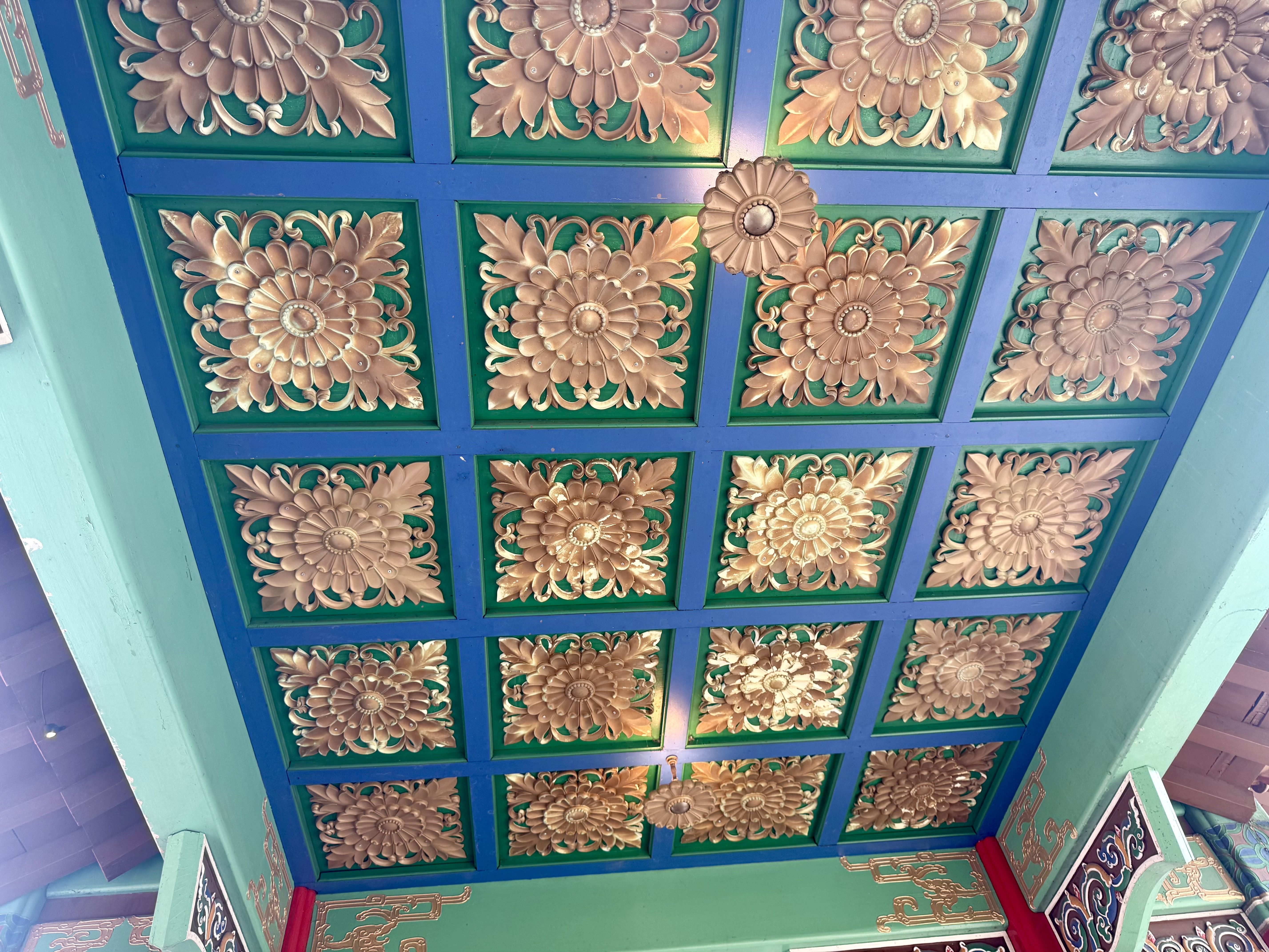 Decorative ceiling panels featuring gold floral rosettes on green backgrounds, framed by blue crossbars; a repeating grid of intricate, daisy-like flowers.
