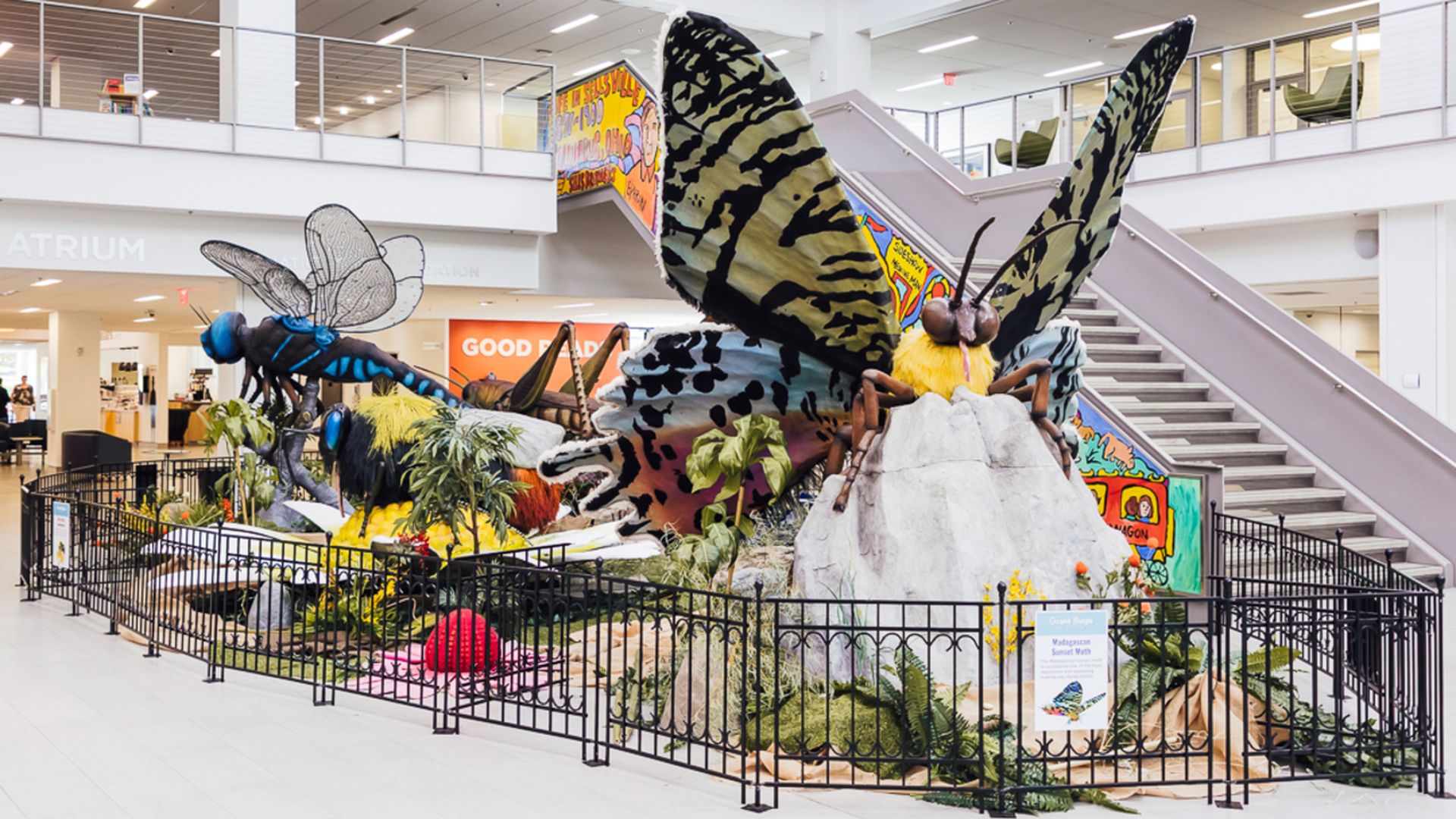 Large indoor display with oversized colorful insect sculptures including a butterfly, bee, and dragonfly, surrounded by plants and black fencing under a white ceiling with stairs nearby.