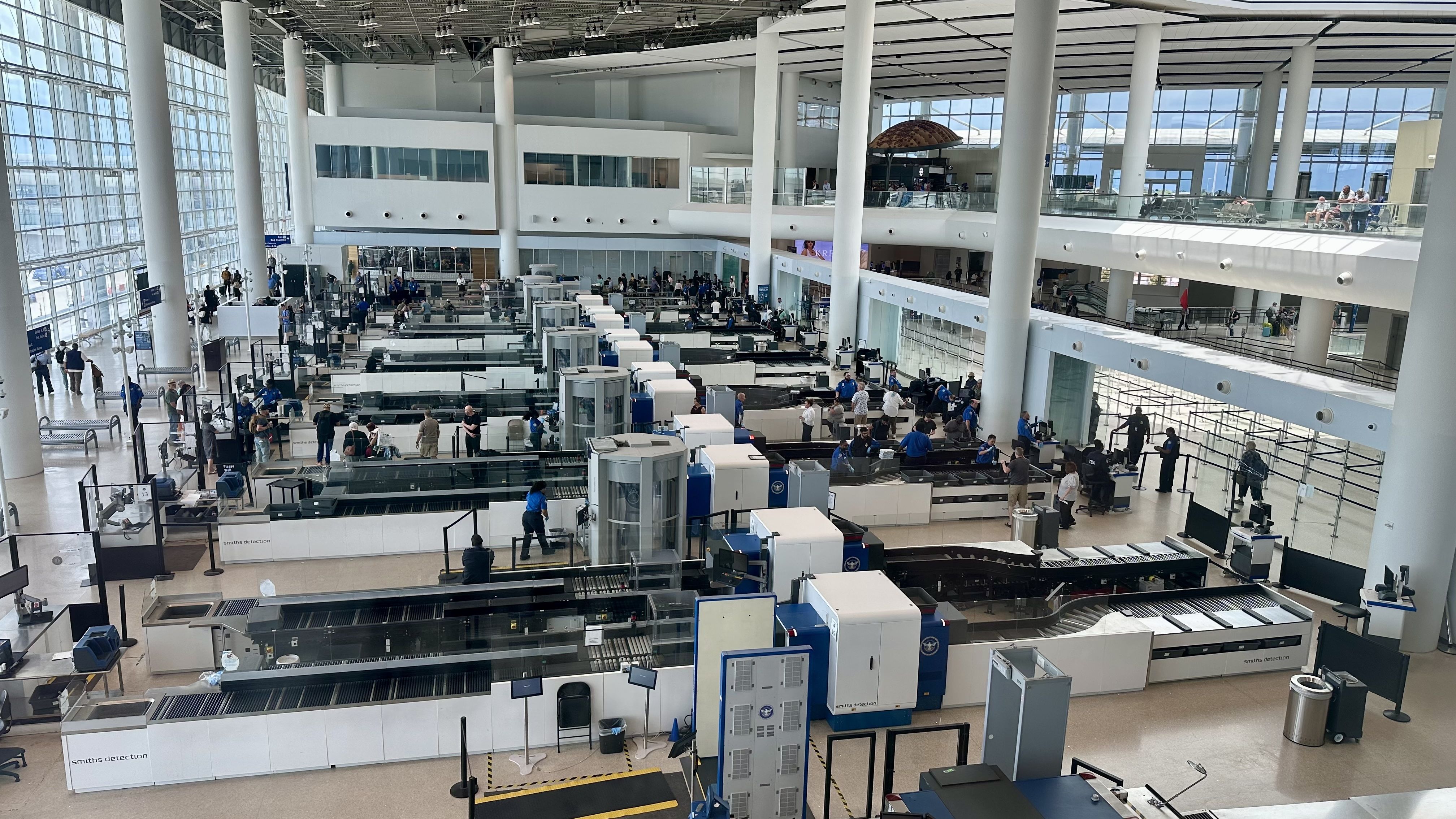 Interior of a large airport security hall with multiple screening belts, travelers in lines, blue-clad staff, white pillars, glass walls, and a mezzanine with seating and shops in daylight.